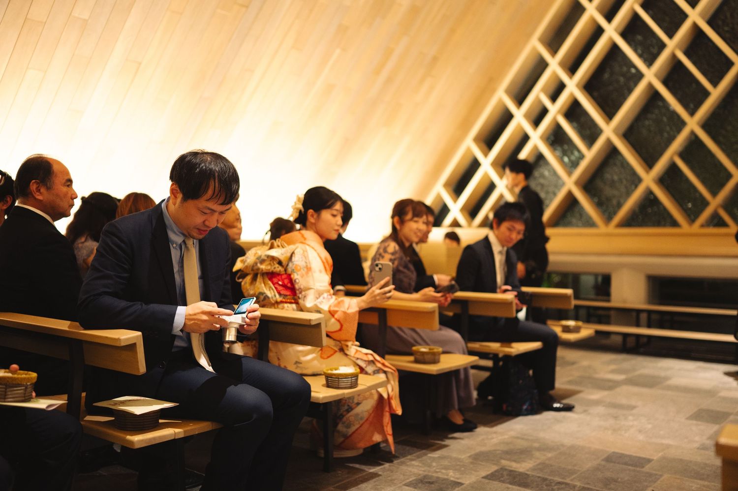 Attendees in formal attire seated in rows at an indoor event with distinctive diagonal lattice ceiling design.