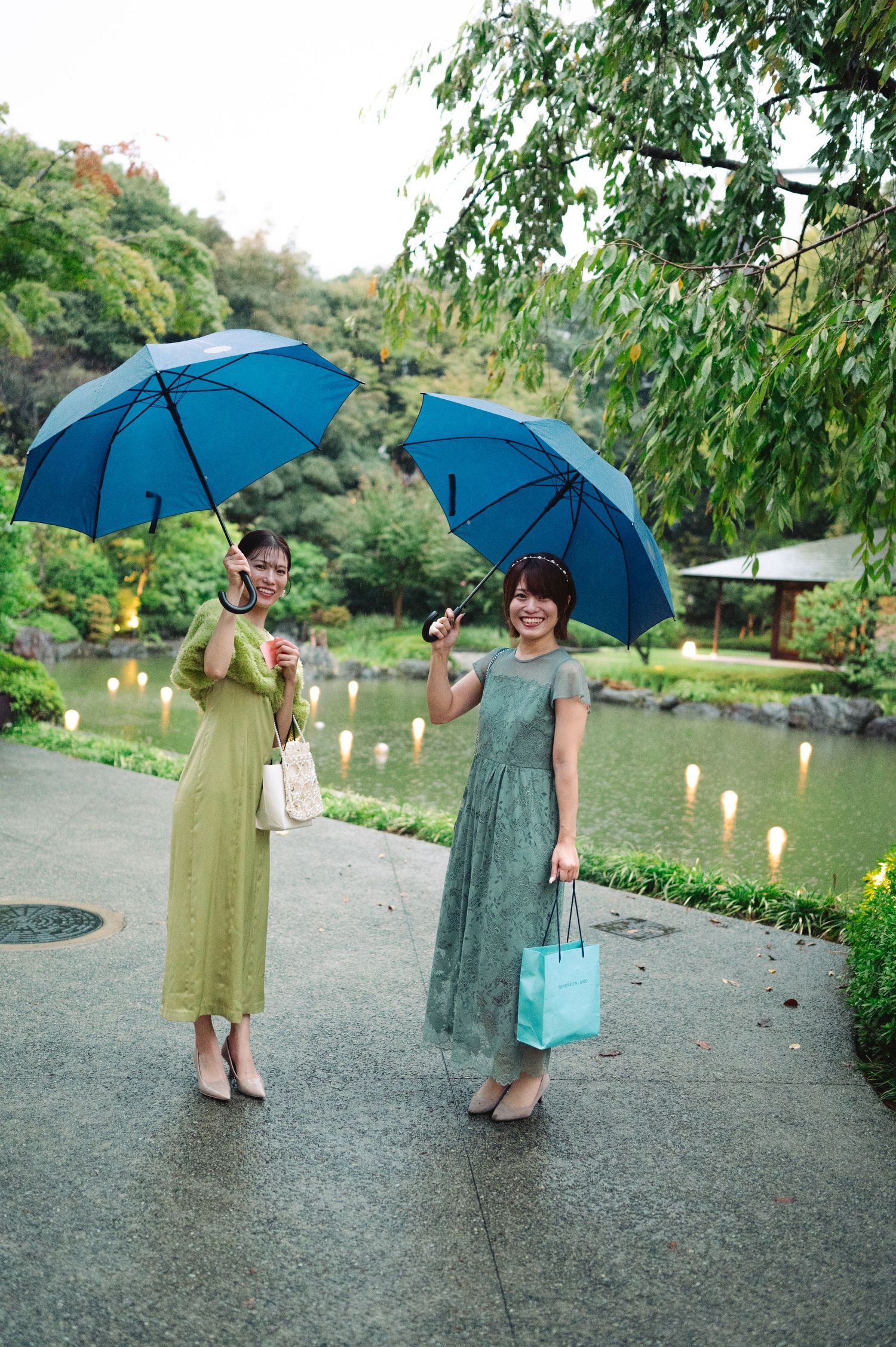 Two people with blue umbrellas walk along a garden path at dusk with lanterns glowing in the background.