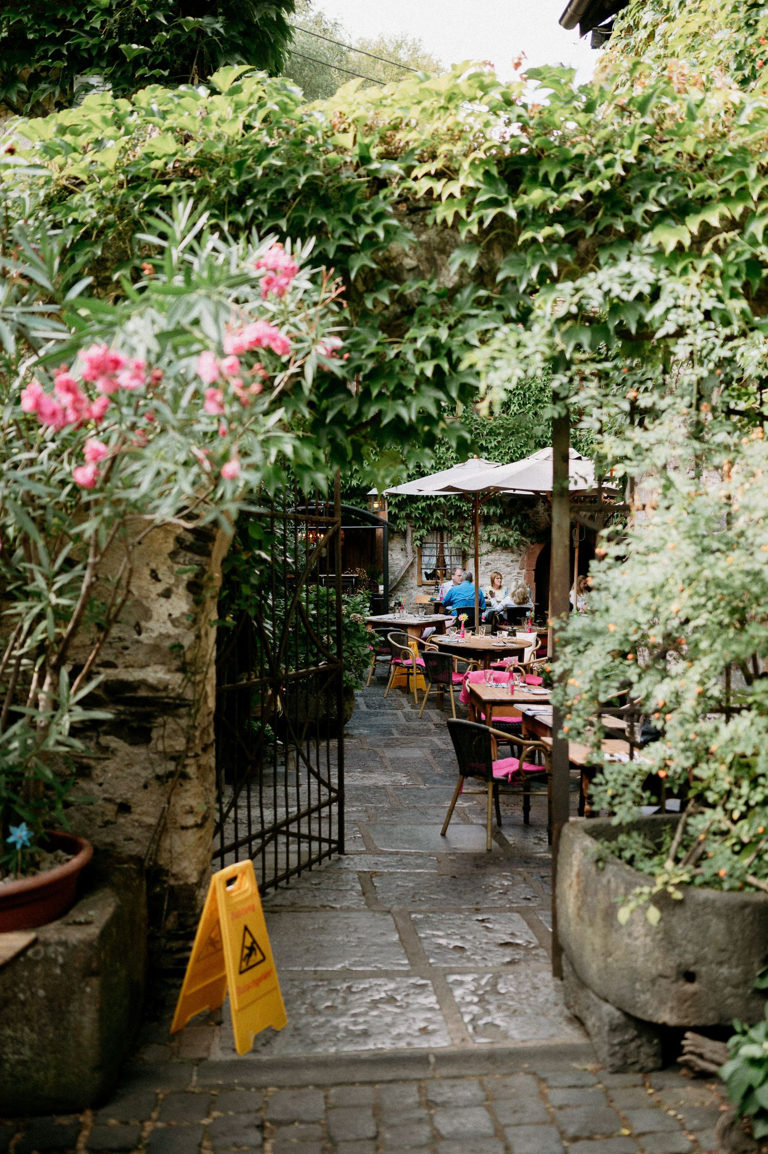 Charming cobblestone courtyard with cafe seating surrounded by flowering plants and stone walls.