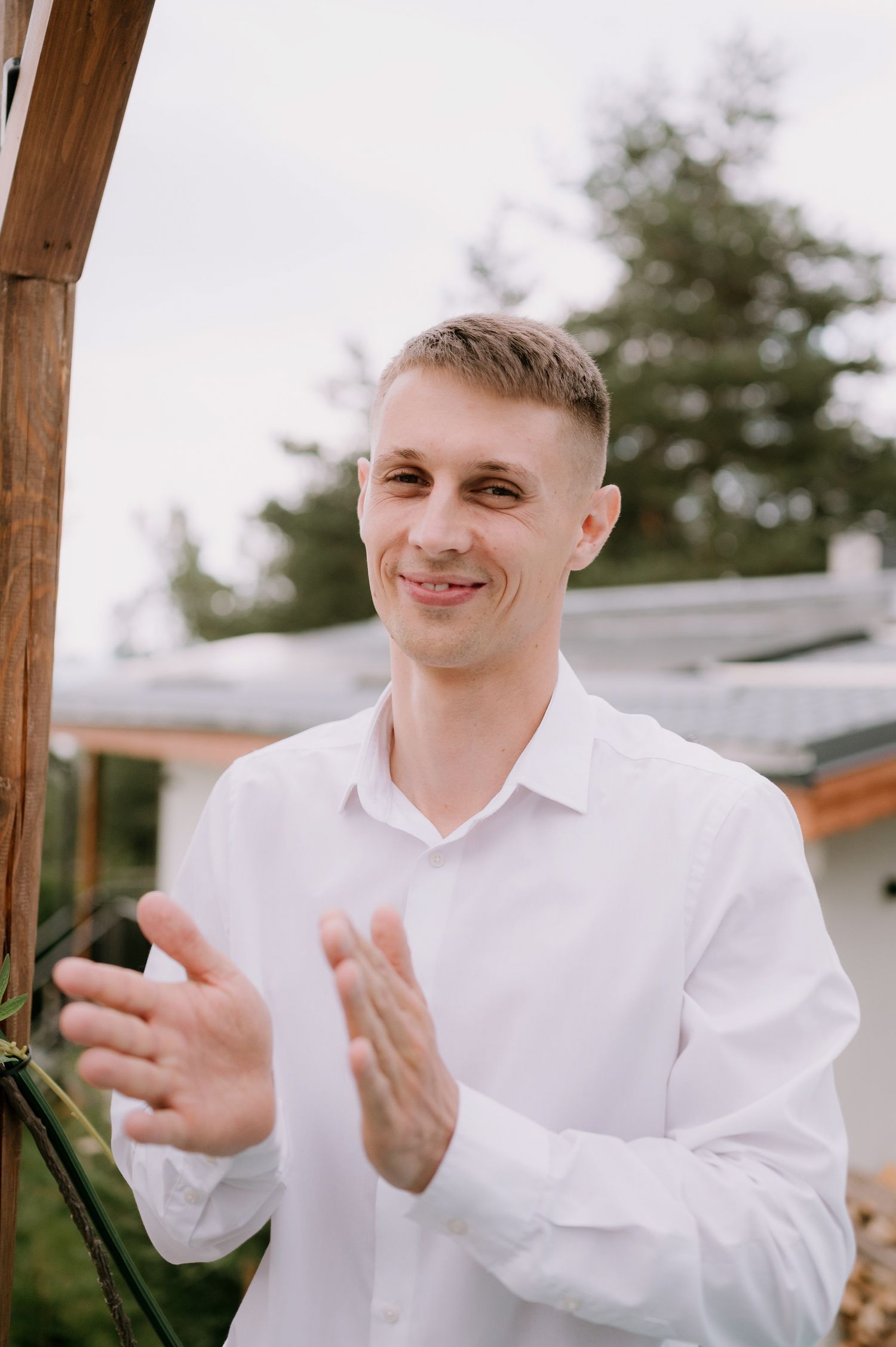 Person in white dress shirt smiling and gesturing outdoors under wooden beams.