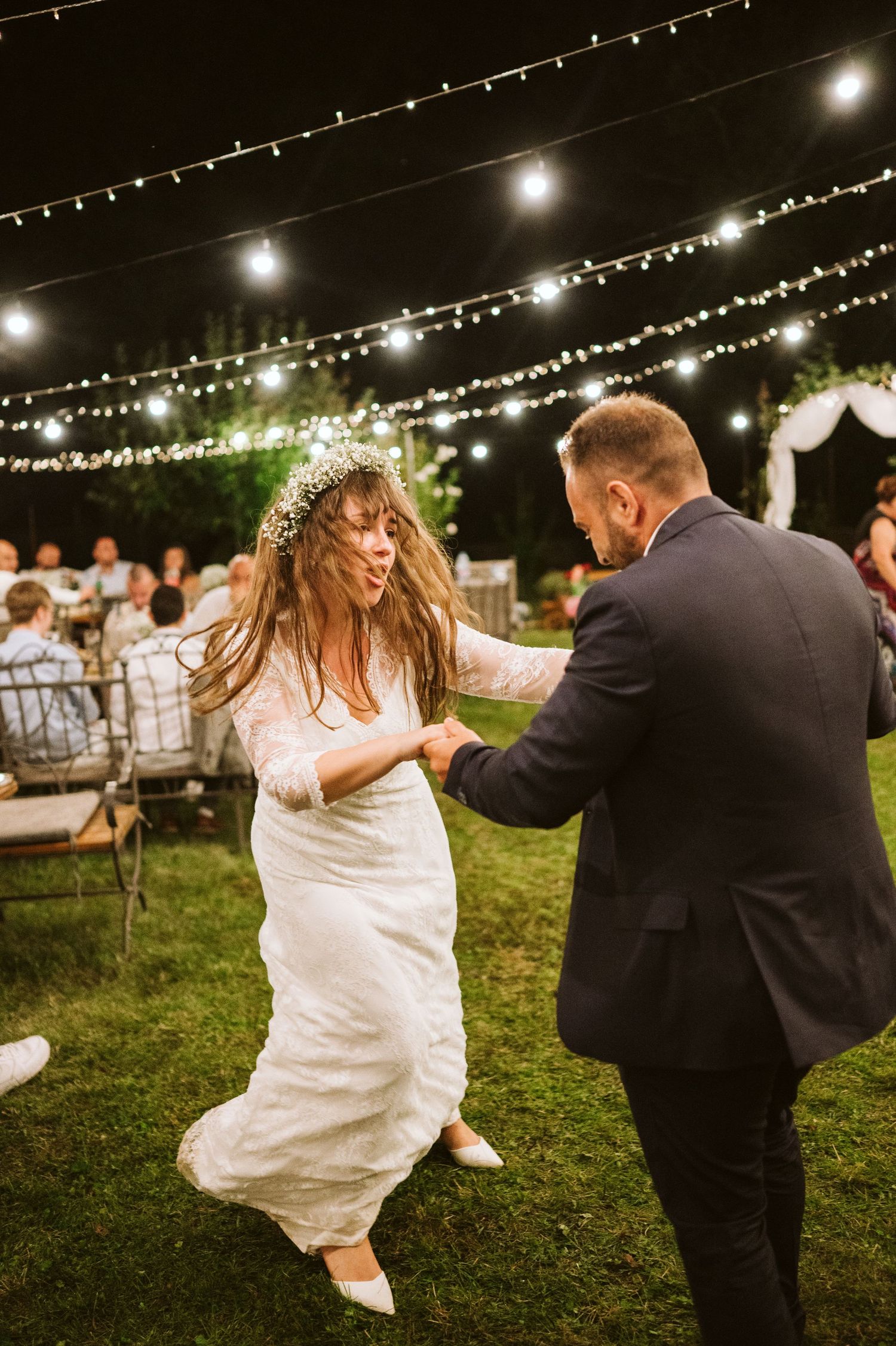 A couple dances joyfully under twinkling string lights at an outdoor wedding reception.