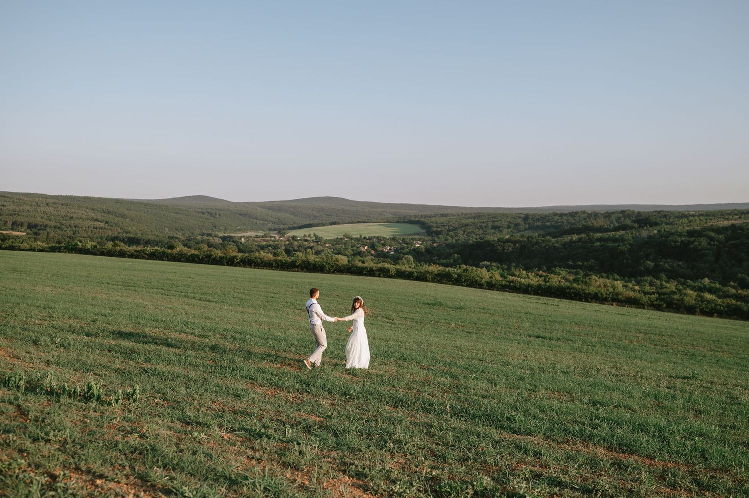 Two figures hold hands while standing in a sweeping green landscape.