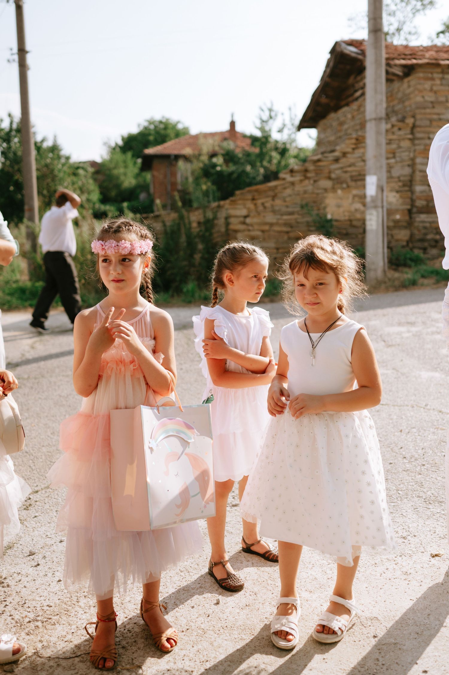 Young flower girls in pink and white dresses walking together on a sunny stone path.