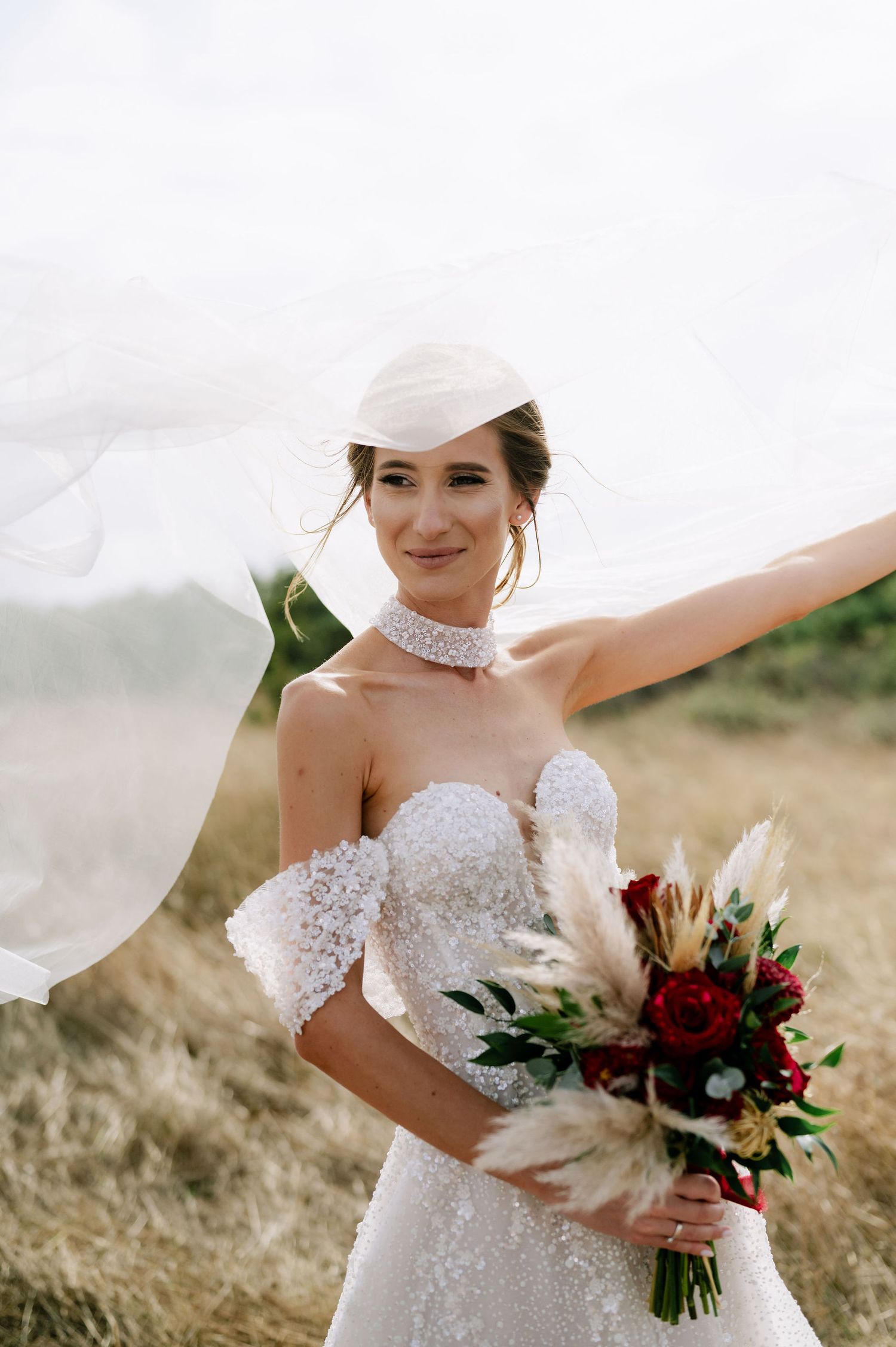 A bride poses with her flowing veil and holds a romantic bouquet featuring red roses and pampas grass.