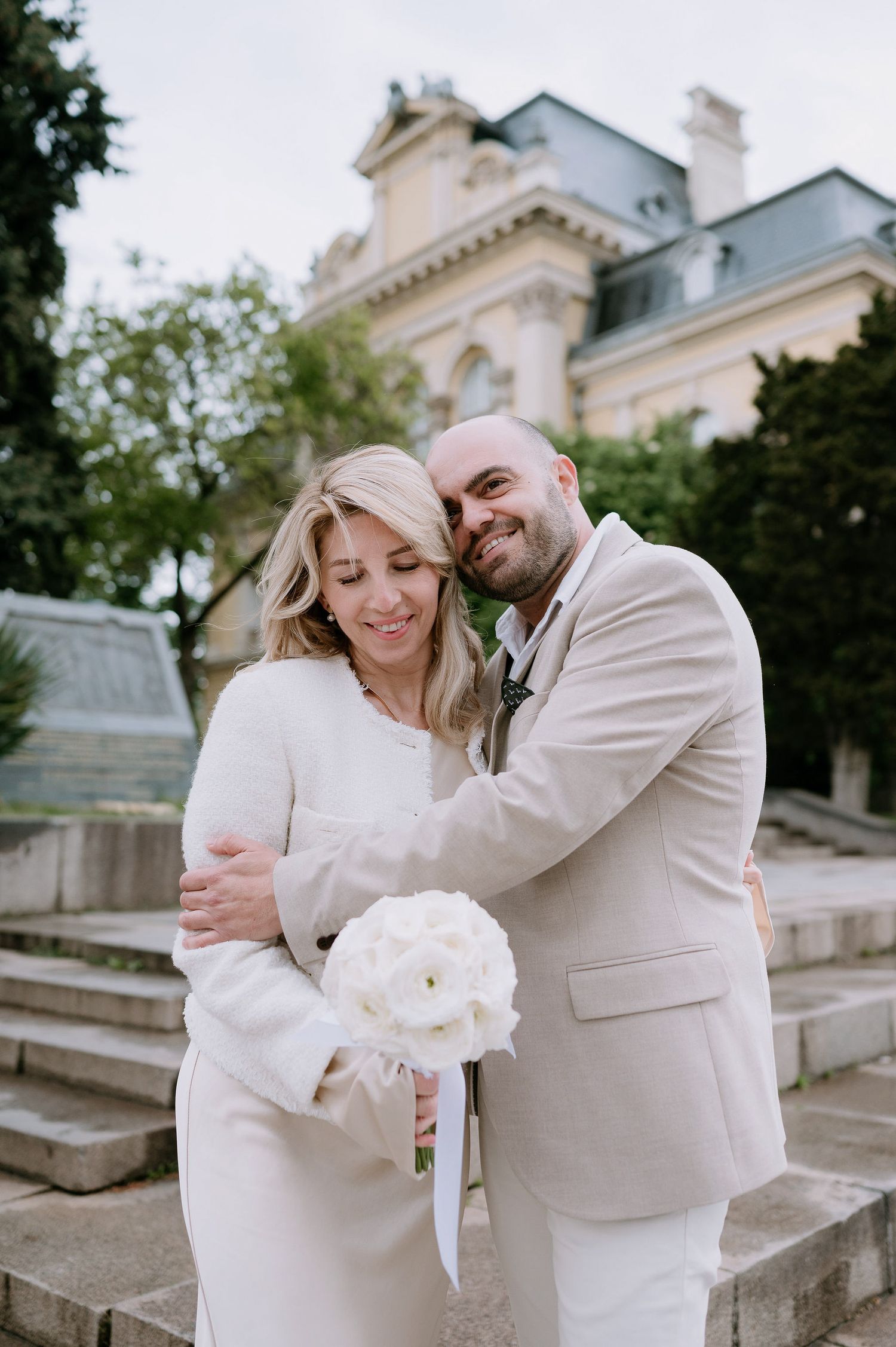 A romantic couple in white embraces while holding a bouquet near stone steps of a historic building.