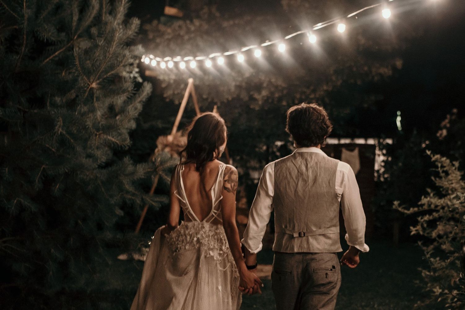 String lights illuminate a romantic outdoor wedding reception celebration at night under the trees.