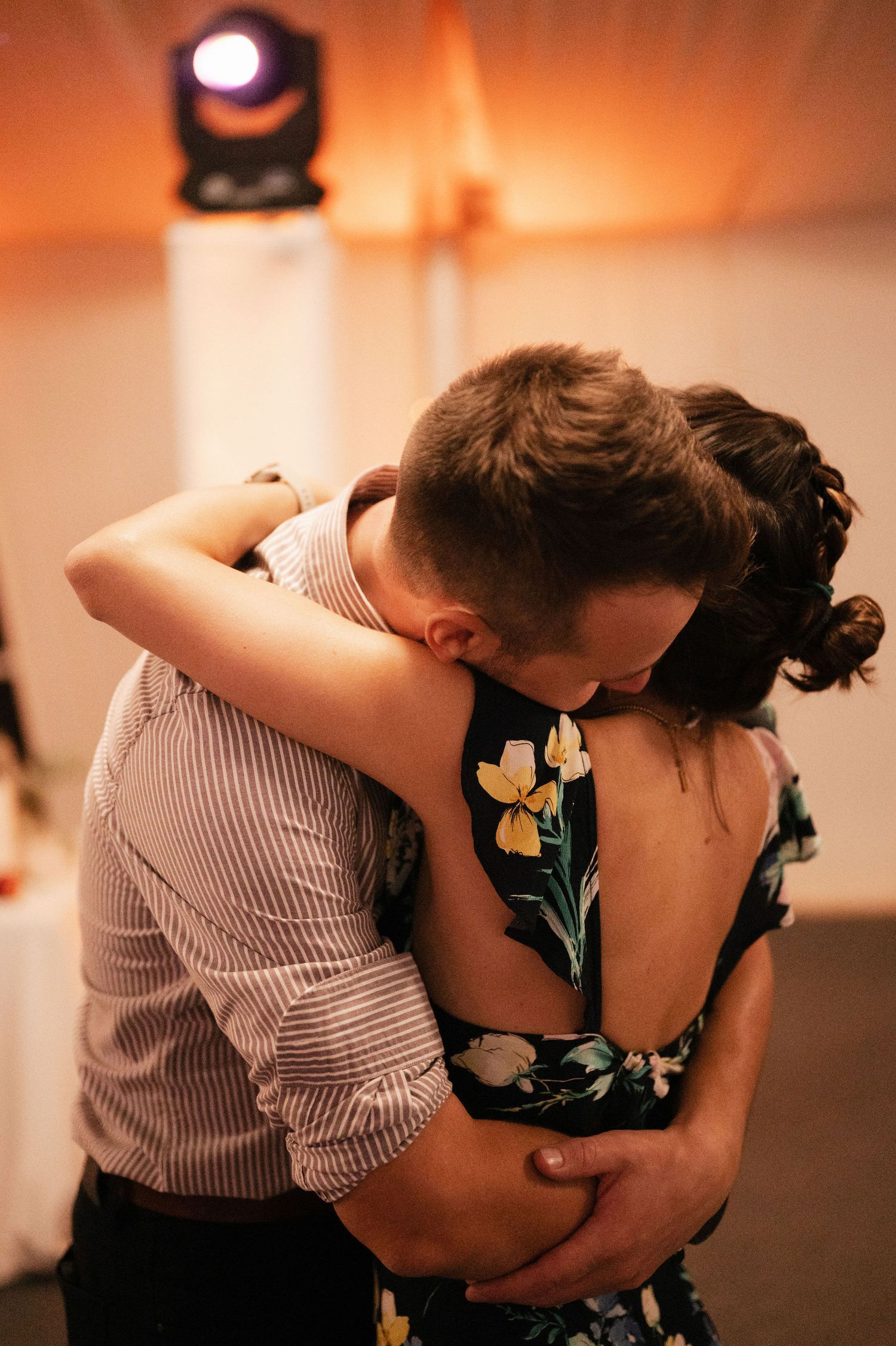 A couple in romantic embrace, with one wearing a floral dress and the other in striped shirt, shown in warm lighting.