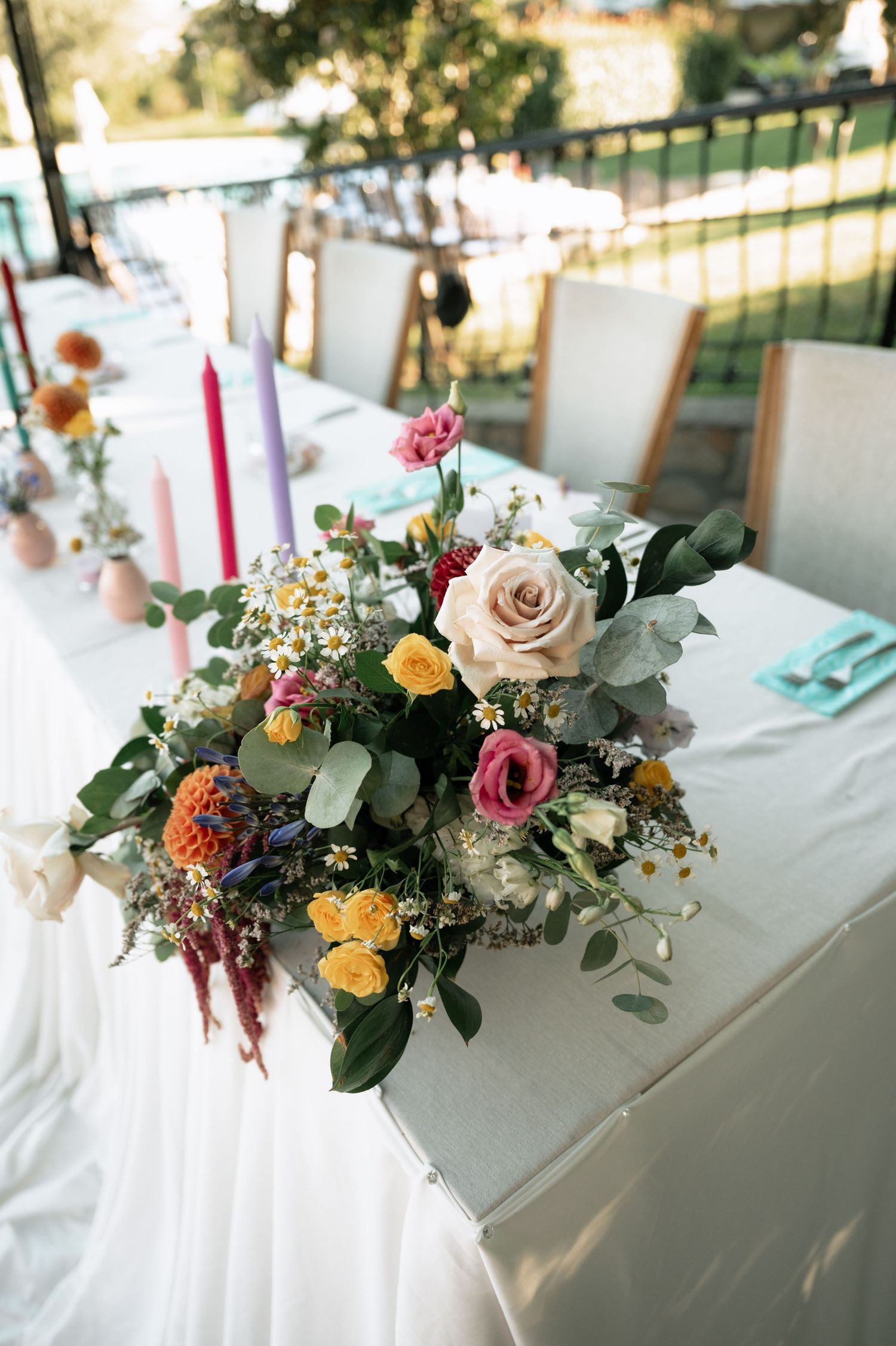A colorful wedding centerpiece with roses, eucalyptus, and mixed flowers on a white tablecloth in an outdoor setting.