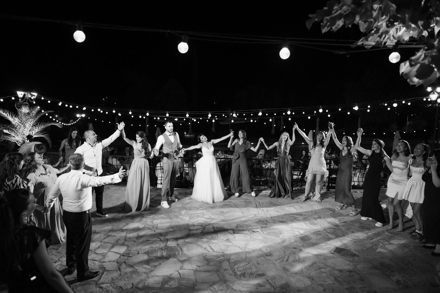 Wedding guests dance together in a circle on an outdoor dance floor under string lights at night during reception celebration.