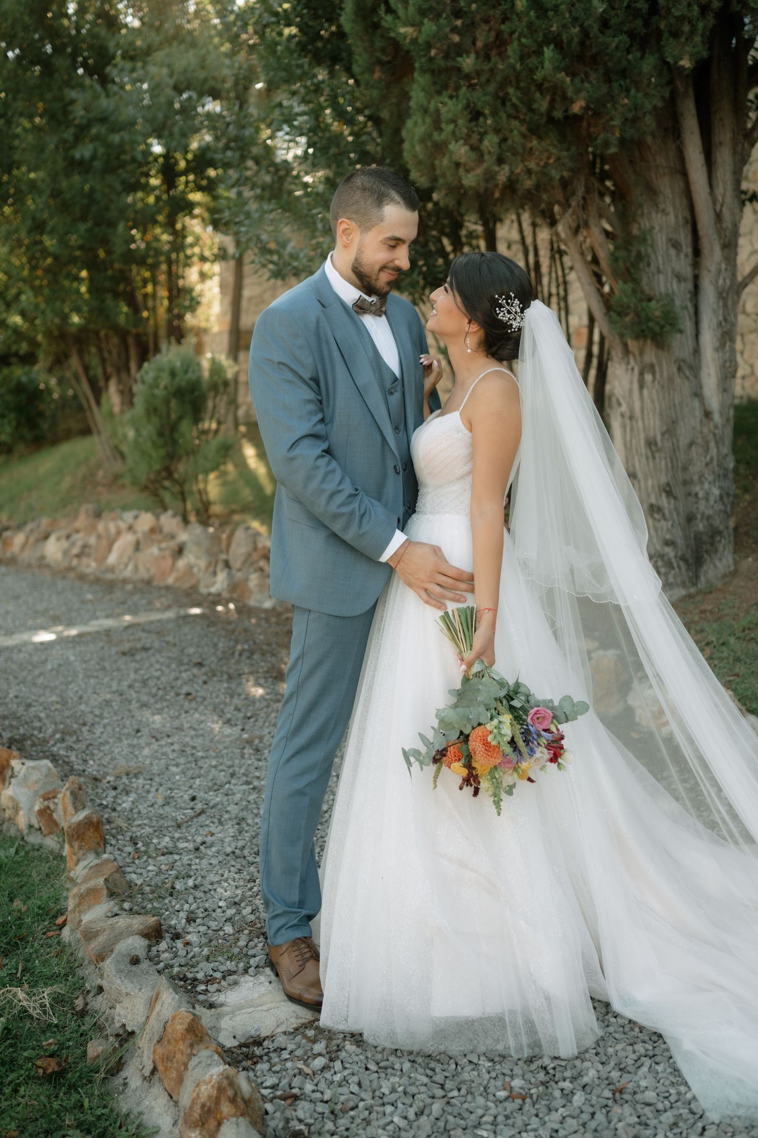 A bride in a white gown with flowing veil holds a coral bouquet while embracing her groom in a gray suit on a garden path.