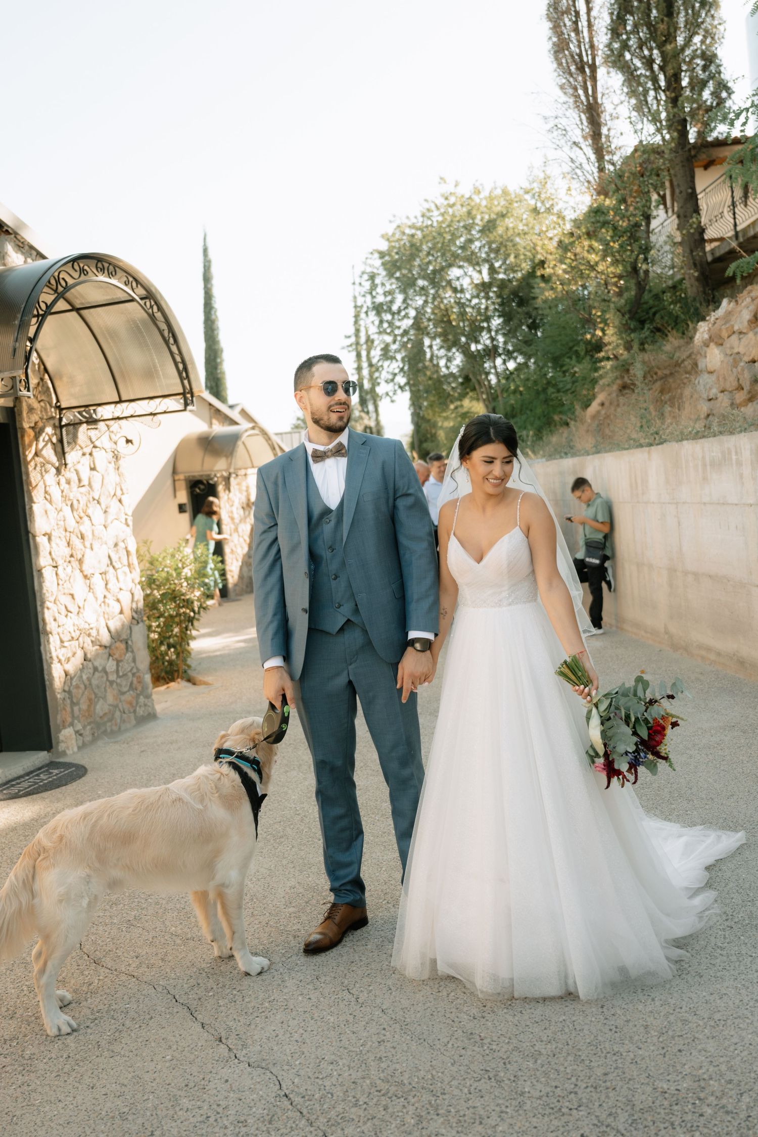 A couple in wedding attire walks down a path while a dog playfully photobombs their romantic moment.