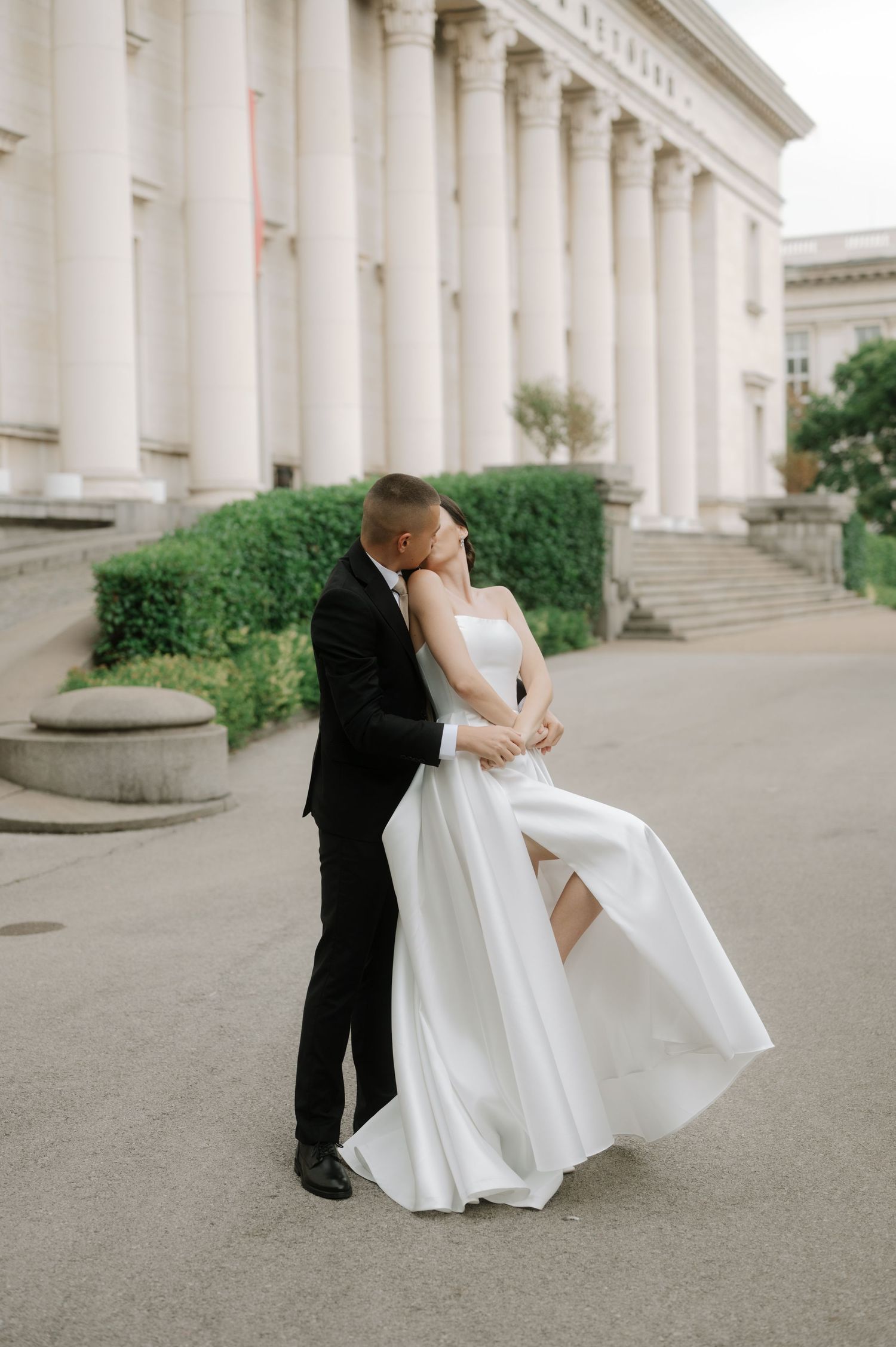 A romantic wedding couple embraces in front of a grand columned building with classic architecture.