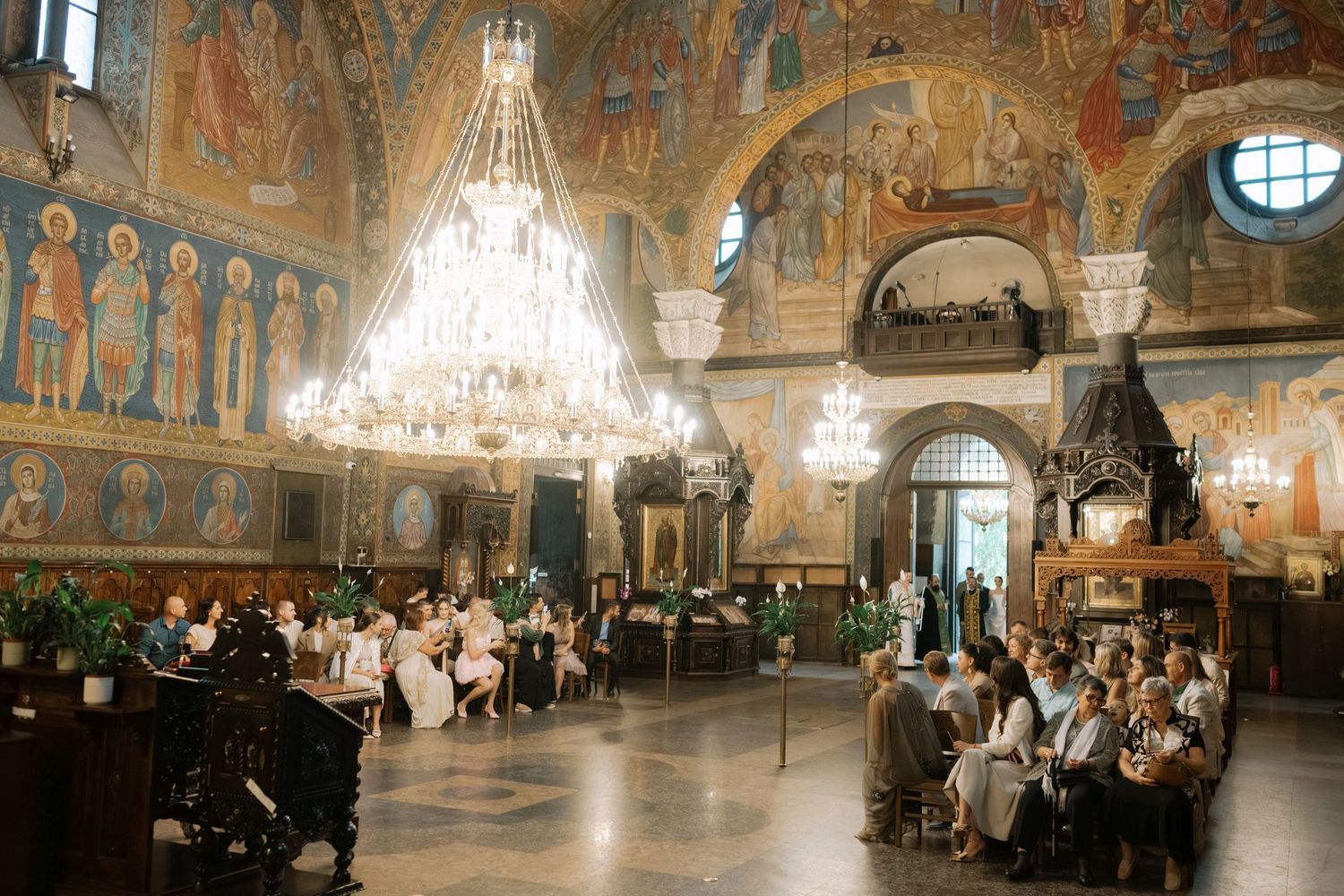 Panoramic view of an ornate palace ballroom with crystal chandeliers, frescoed walls, and visitors exploring the grand space.