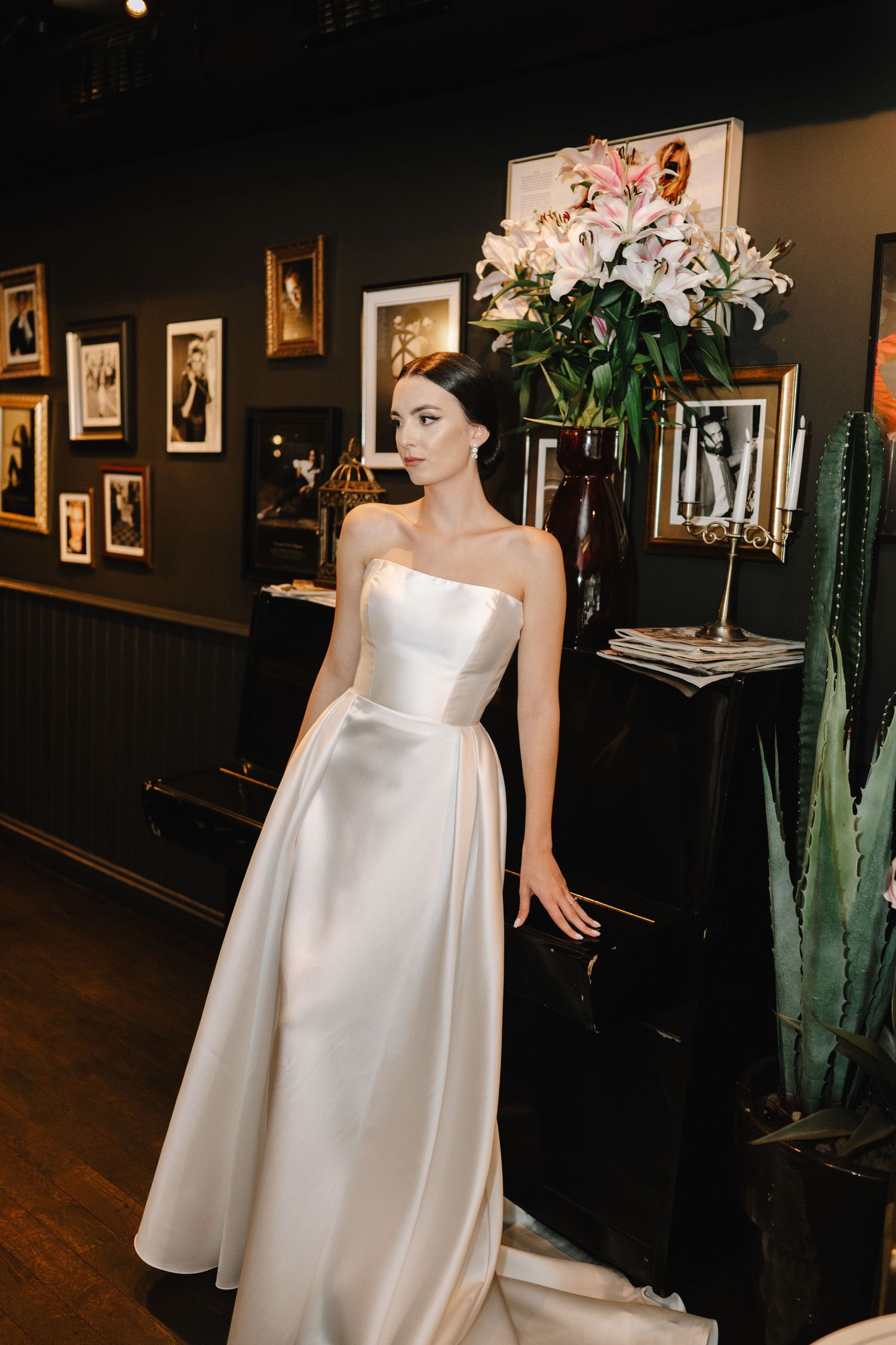 A white wedding dress with a flowing train is displayed in a dark gallery space with artwork and cactus plants.