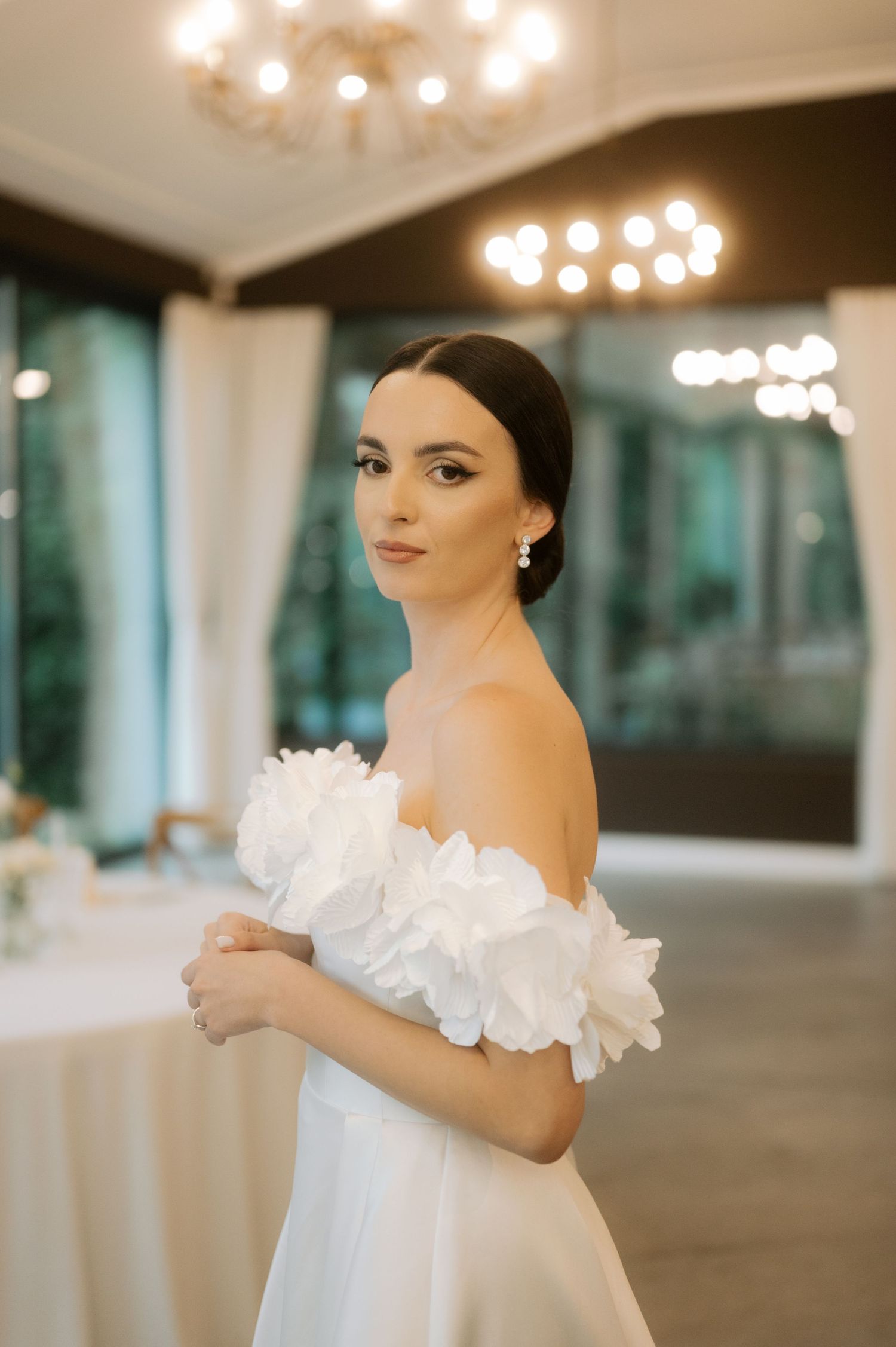 A bride in an elegant white off-shoulder wedding gown poses under glowing chandeliers in an elegant venue.