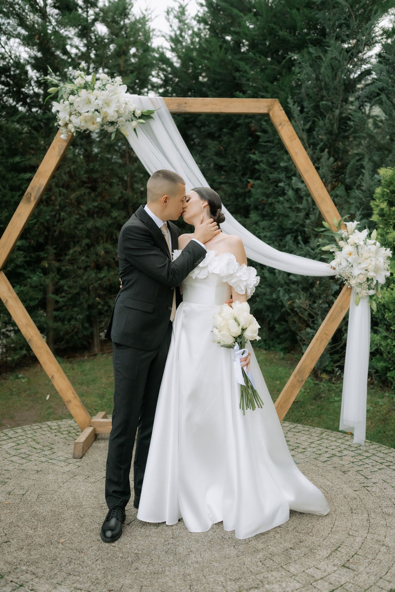Newlywed couple shares romantic kisses in front of a wooden geometric ceremony arch draped with white fabric outdoors.