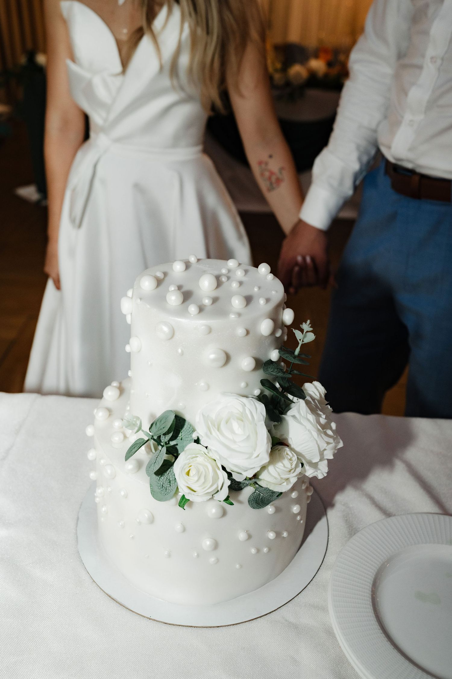 White wedding cake decorated with delicate white roses and pearl accents sits elegantly on display at reception.