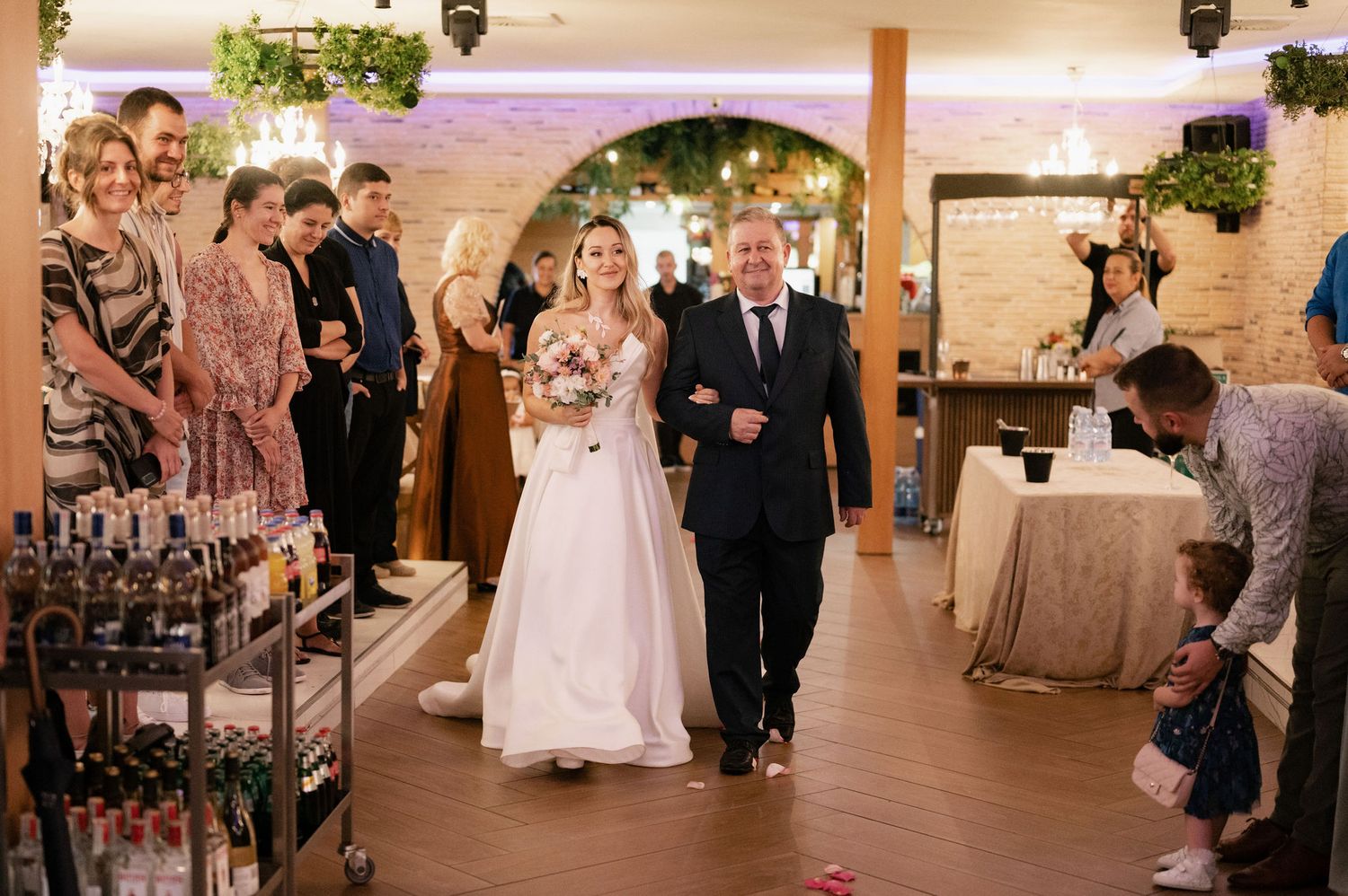 Wedding ceremony procession walking down aisle in rustic brick venue with hanging lanterns and floral decor.