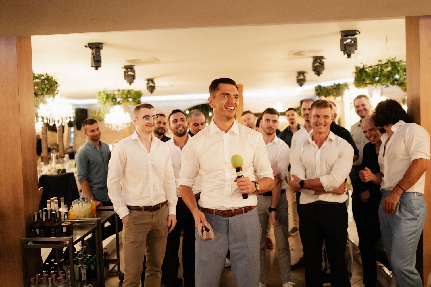 Wedding guests in white shirts and formal attire gather at an indoor reception venue during a celebration.