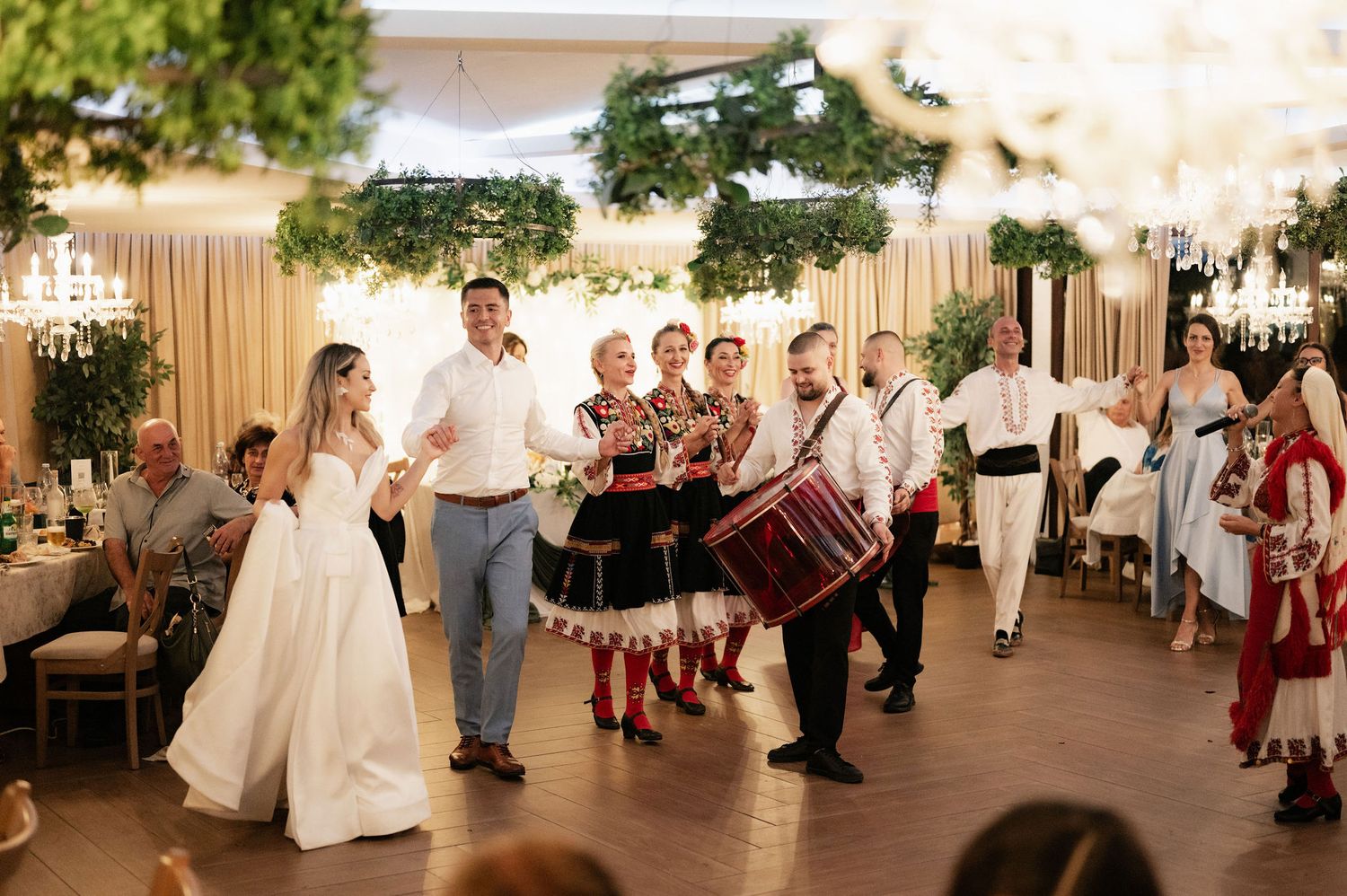Wedding party dances in celebration with traditional folk dancers in red and white costumes performing at reception.