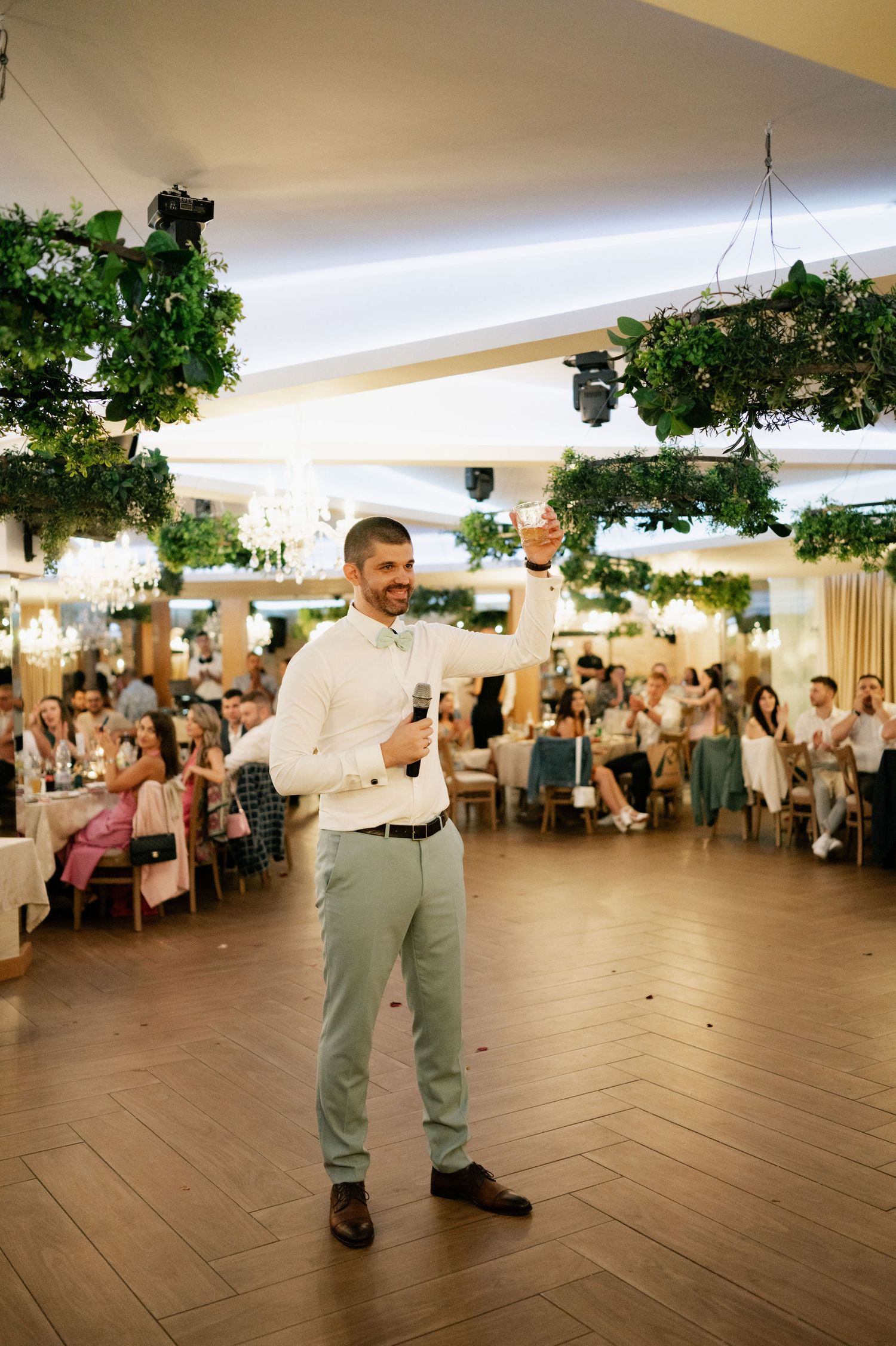 A speaker addresses guests at an indoor wedding reception venue decorated with hanging greenery and ambient lighting.