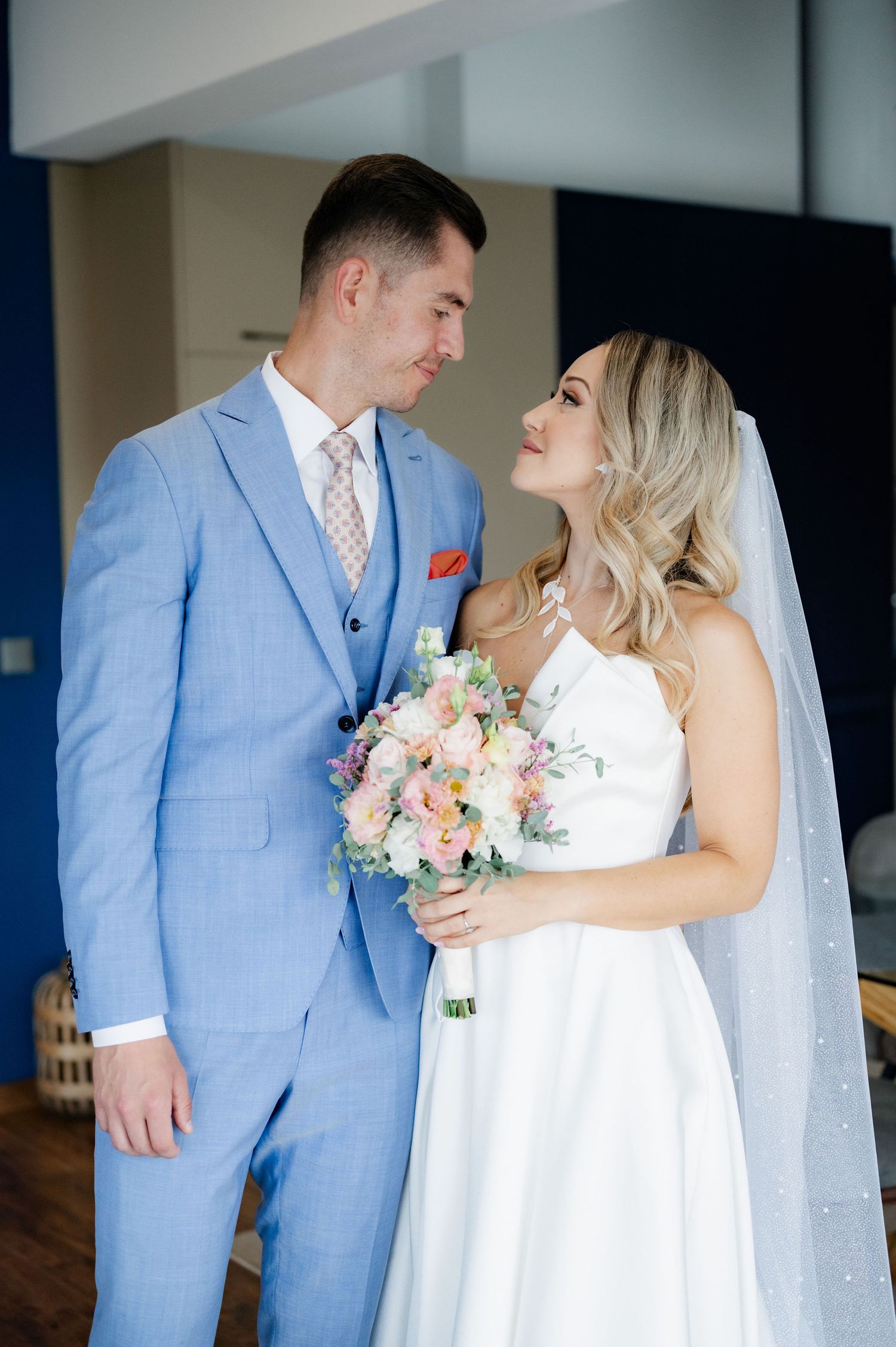 A bride in a white gown holds a pastel bouquet while looking lovingly at her groom wearing a light blue suit.