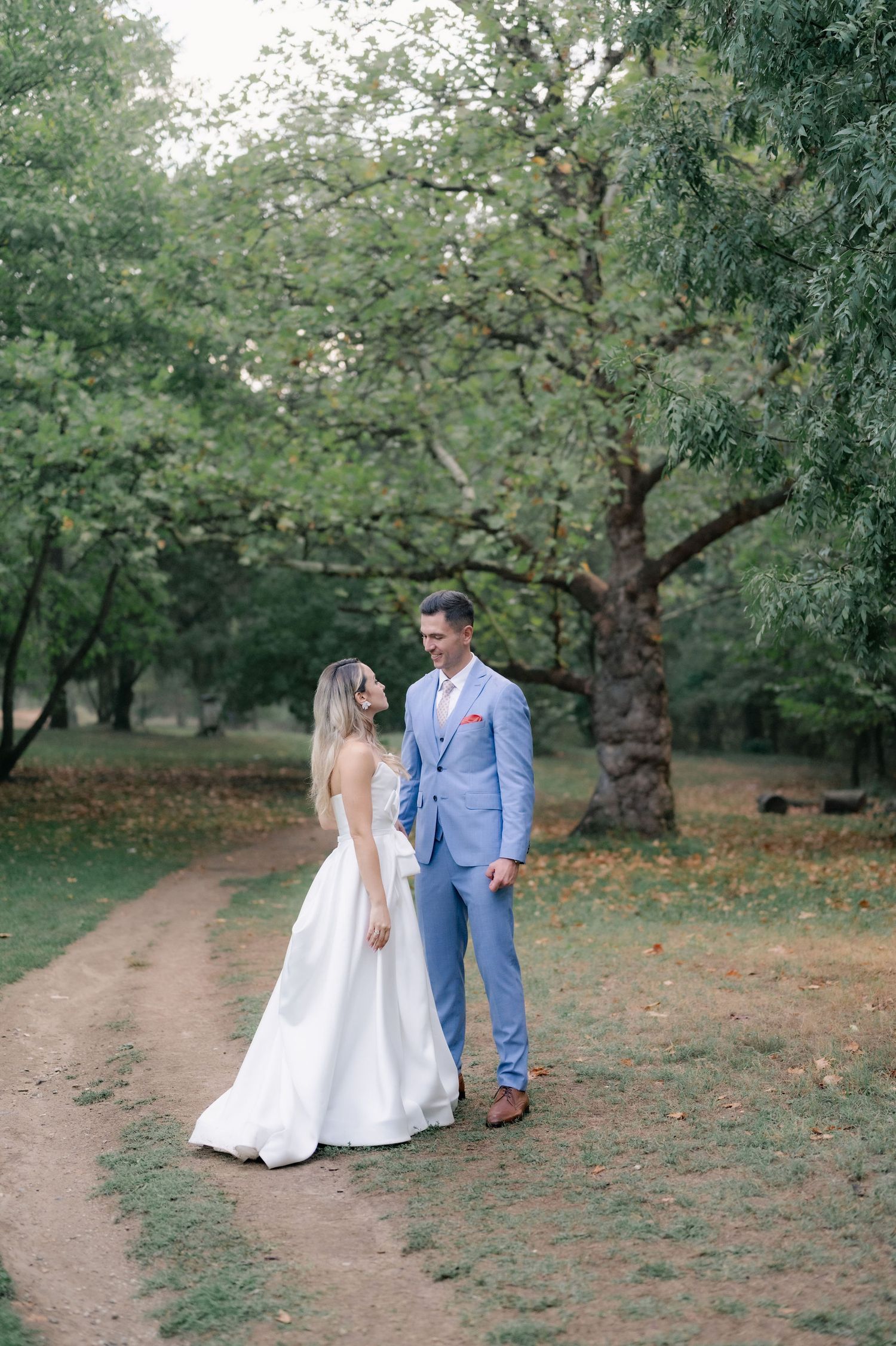 A couple in wedding attire walks down a dirt path surrounded by lush green trees in an orchard setting.