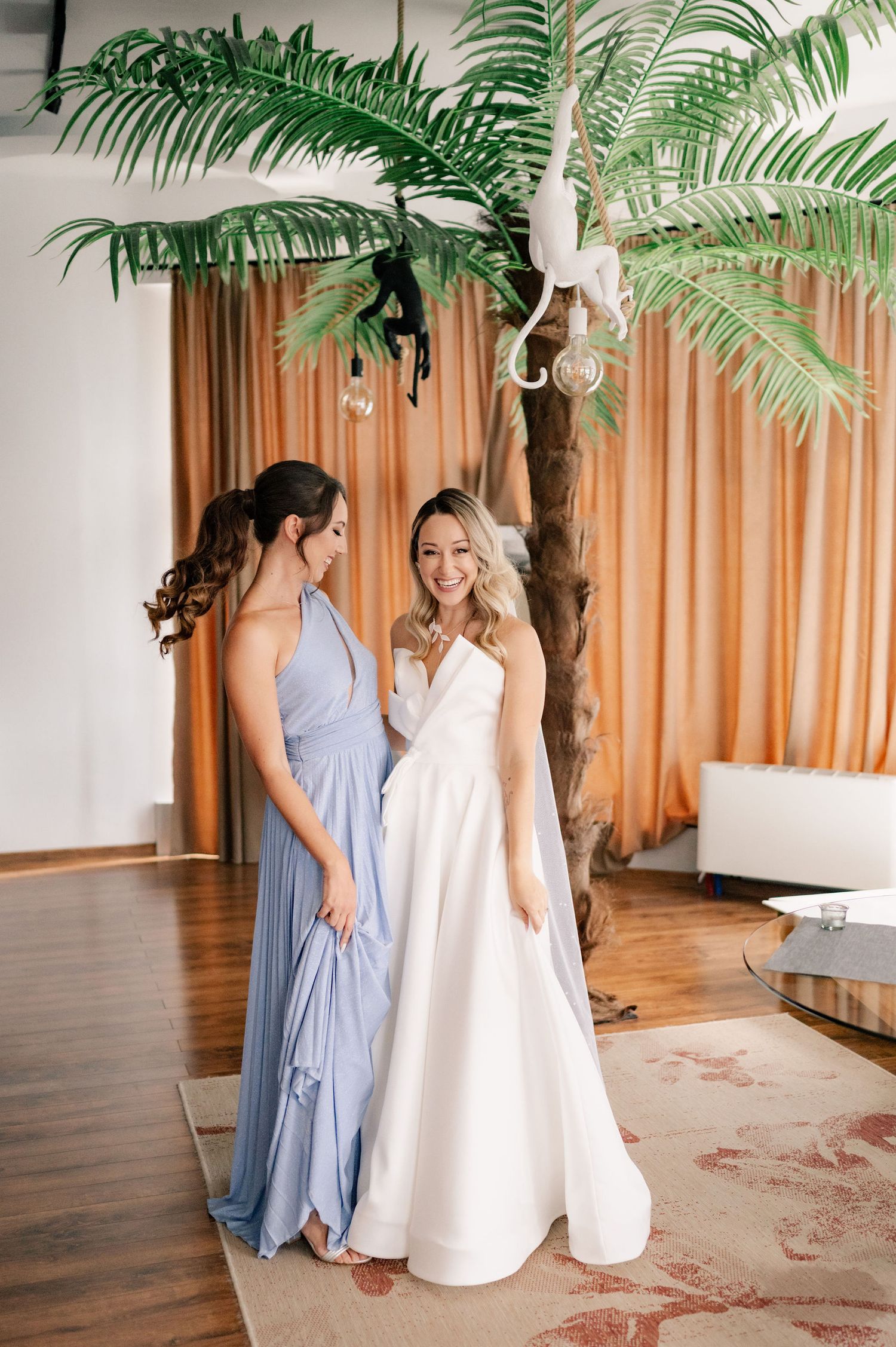 Two women stand together under a palm tree, one wearing a light blue gown and one in a white wedding dress.
