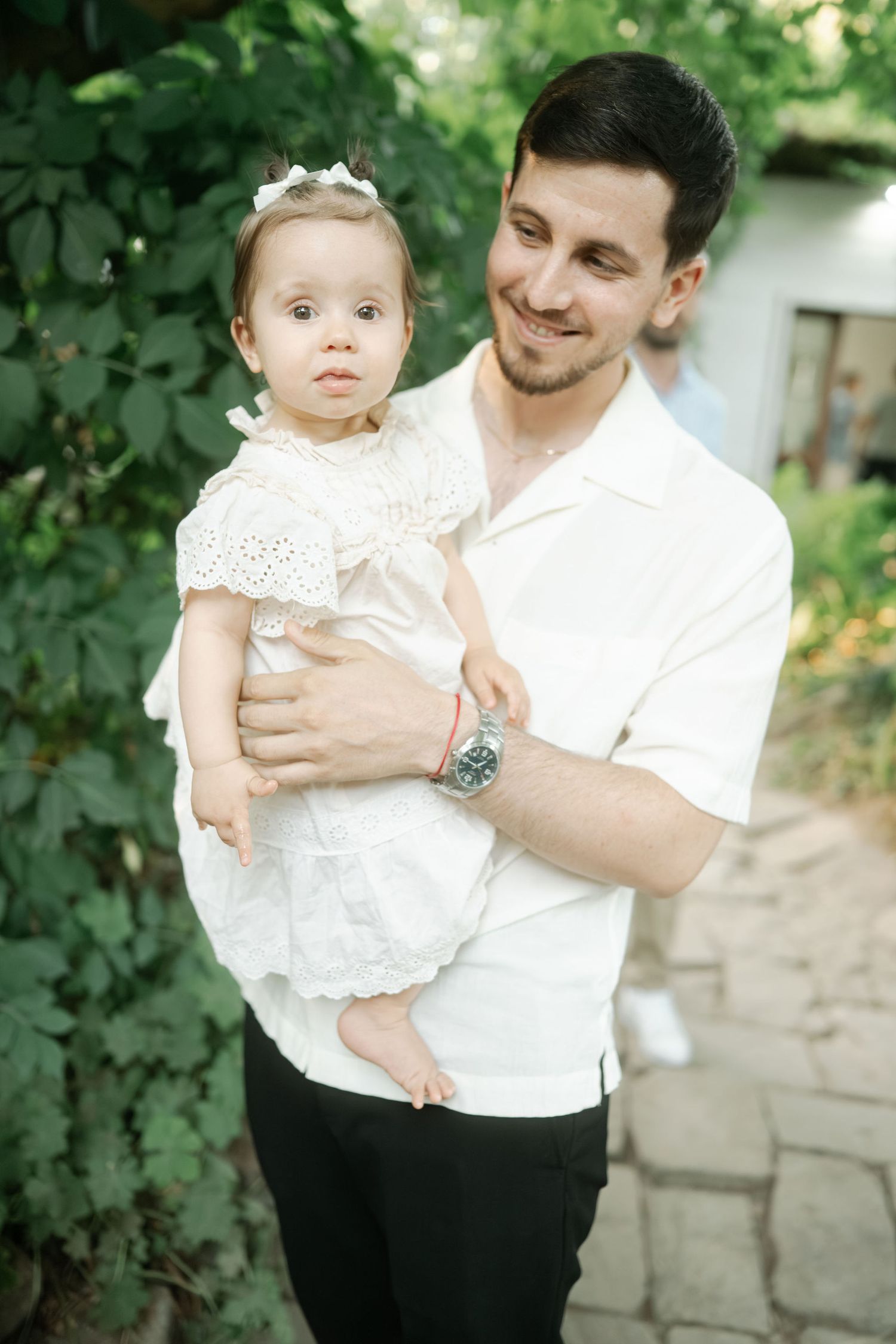 A parent in a white shirt holds a baby wearing a white lace dress in a garden setting.