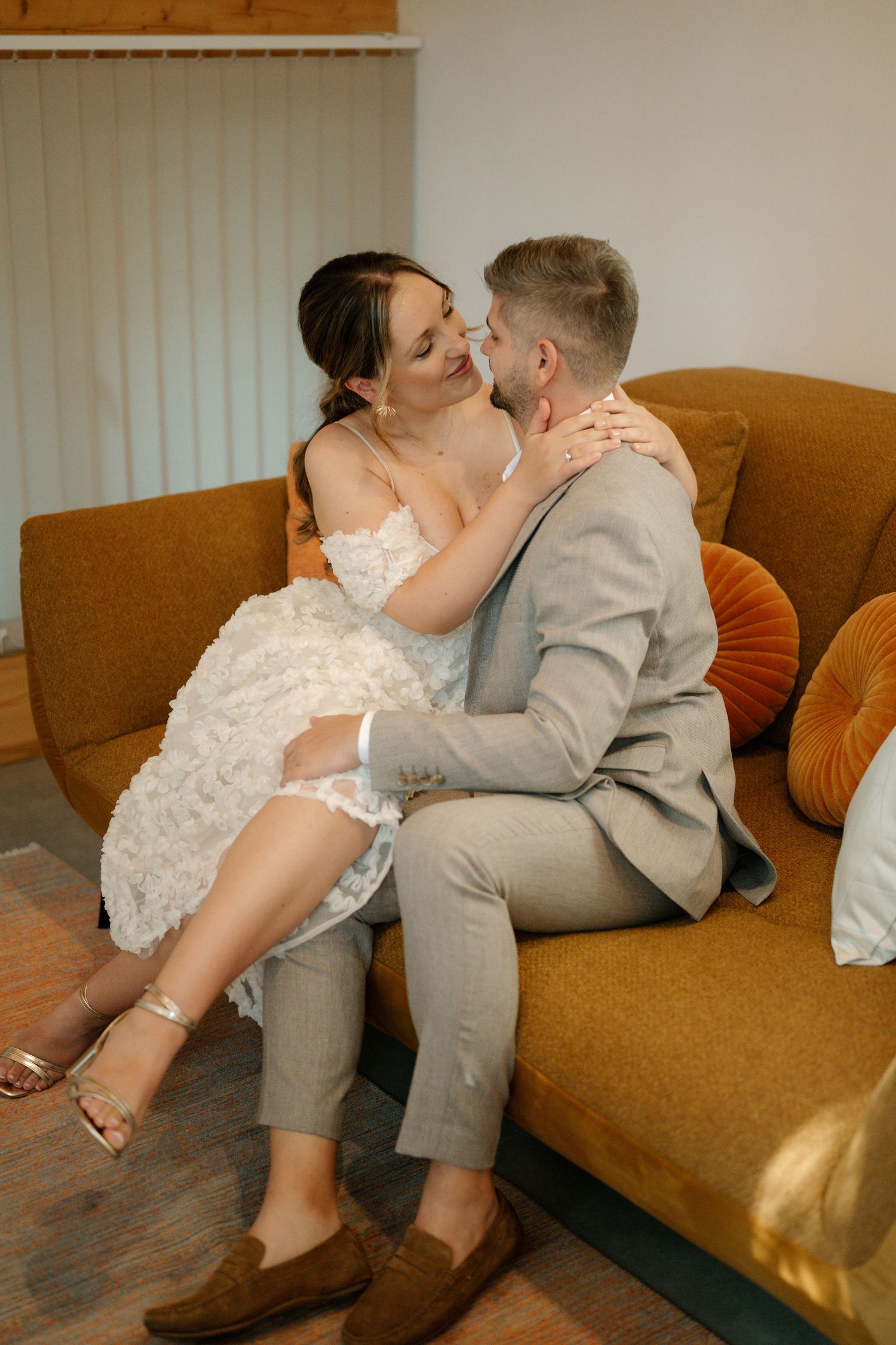 Newlyweds share tender moments on a brown couch in an indoor setting with natural lighting.
