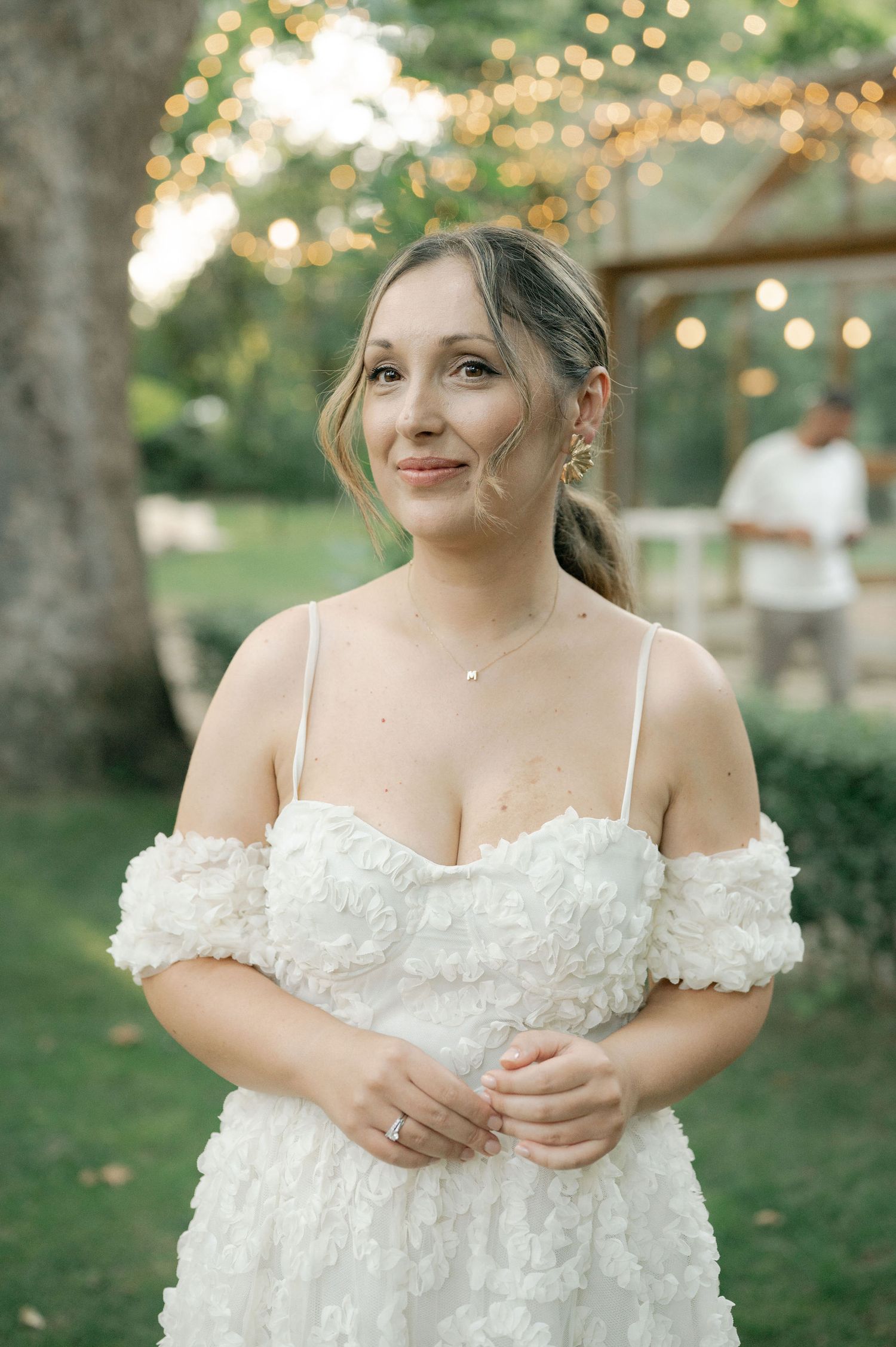 A bride in an off-shoulder white wedding dress poses outdoors with string lights and trees in the background.