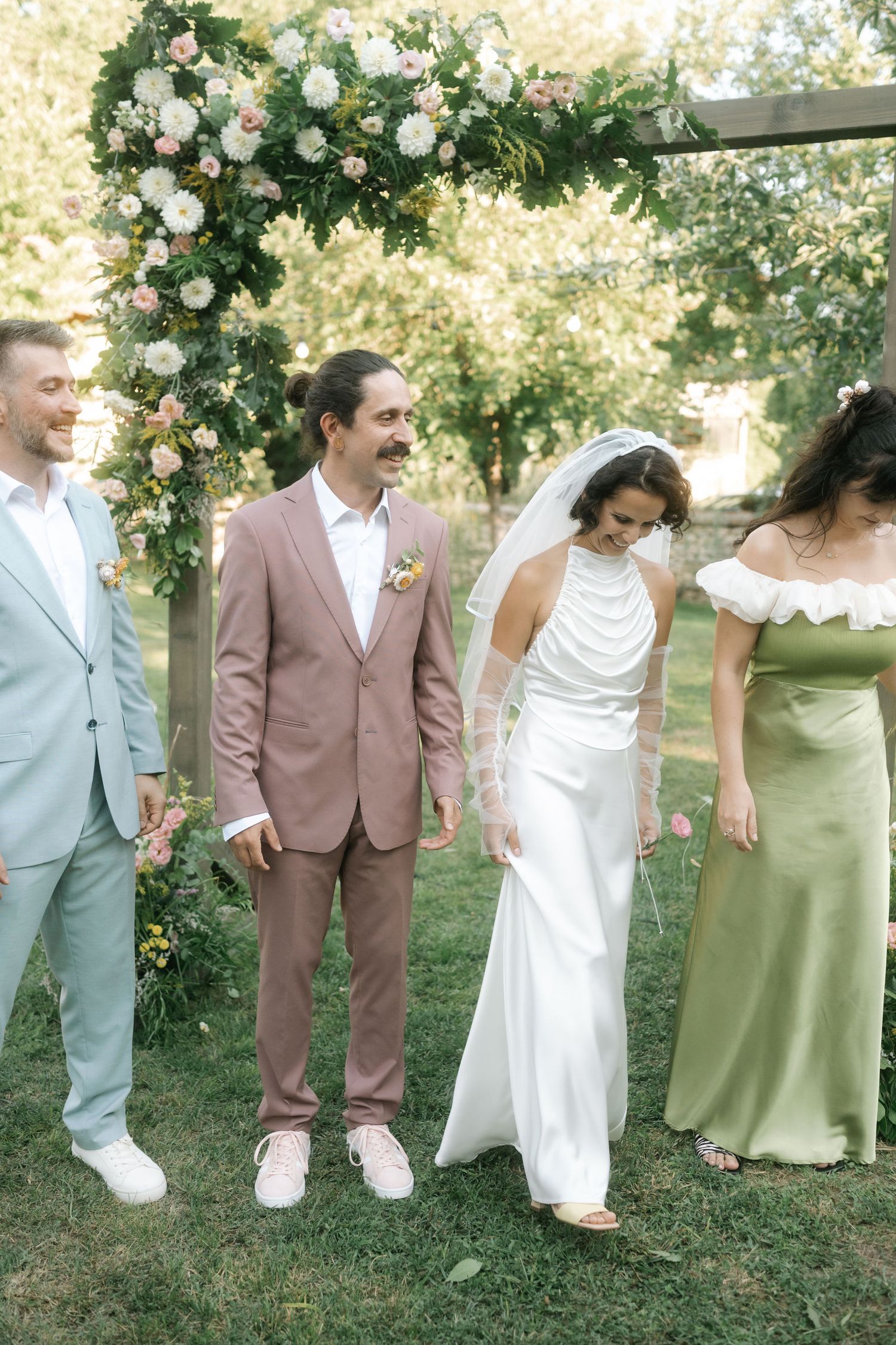 Wedding party members in pastel suits and dresses pose together in front of white floral decorations in an outdoor setting.