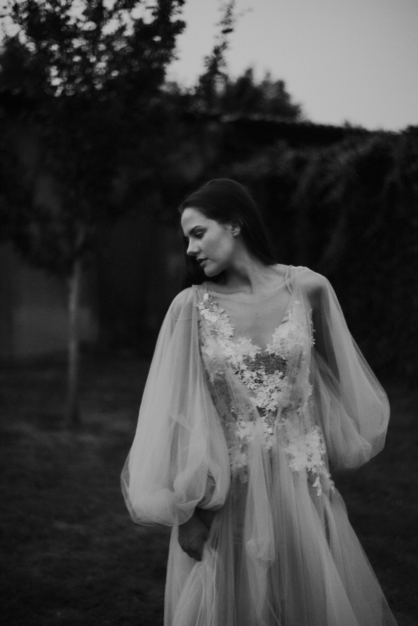 Ethereal black and white photo of a flowing white lace dress with sheer sleeves against a moody forest backdrop.
