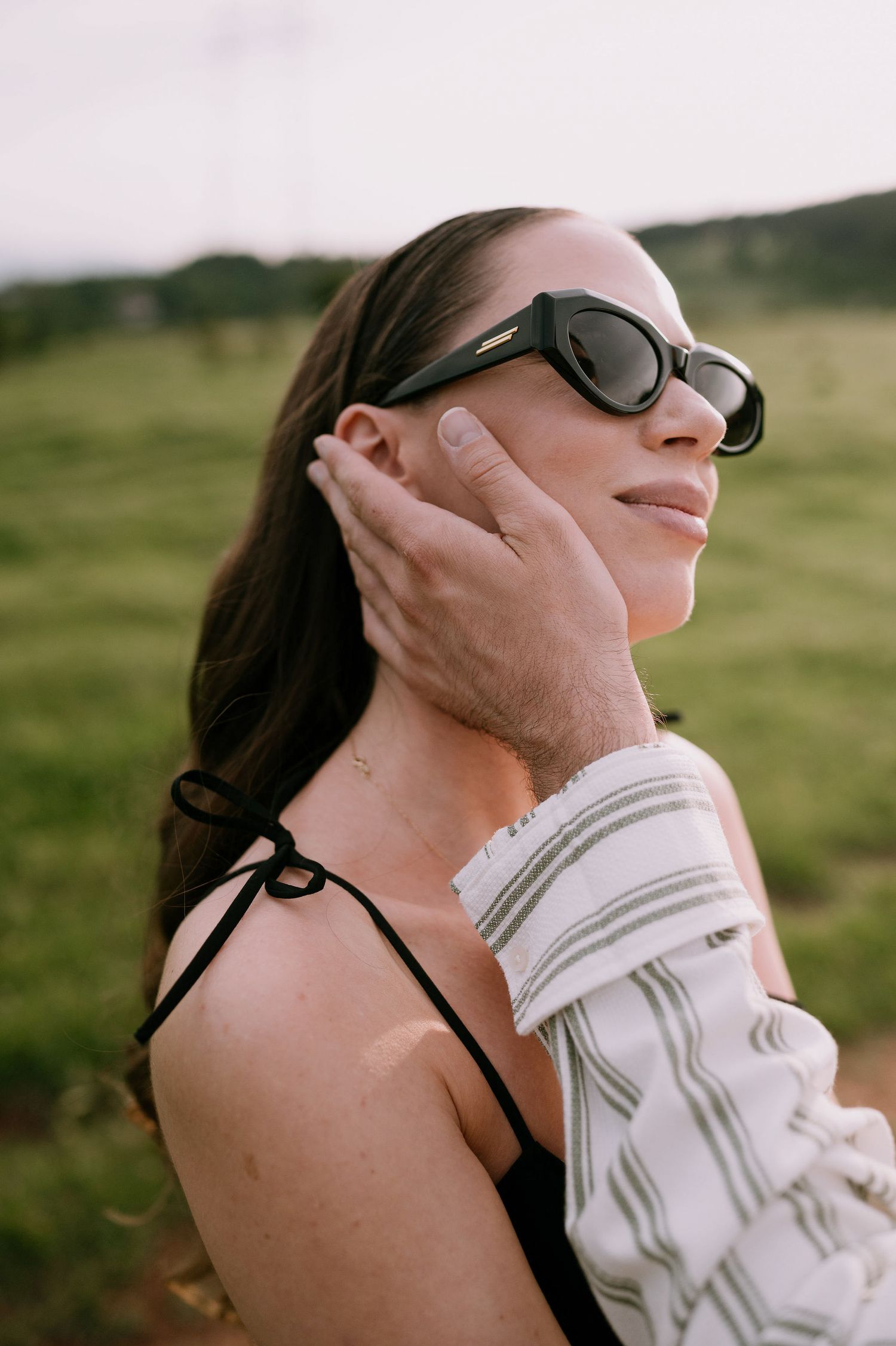 A person wearing sunglasses tilts their head back while wrapped in a white blanket outdoors in nature.