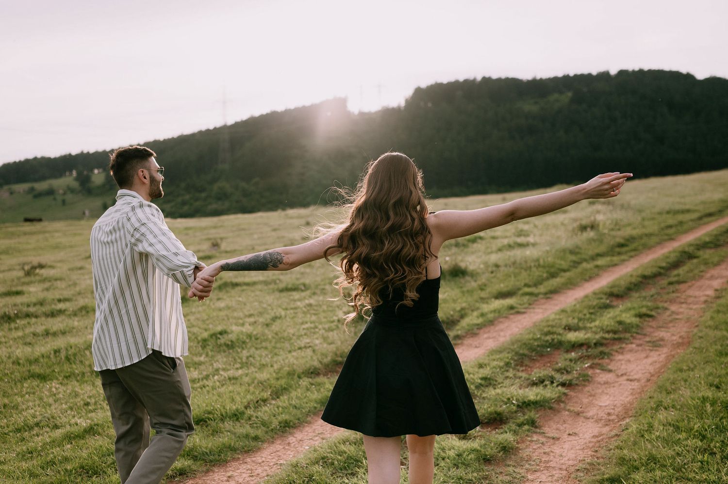 Two people hold hands while walking along a dirt path in a sunny meadow with mountains in the background.