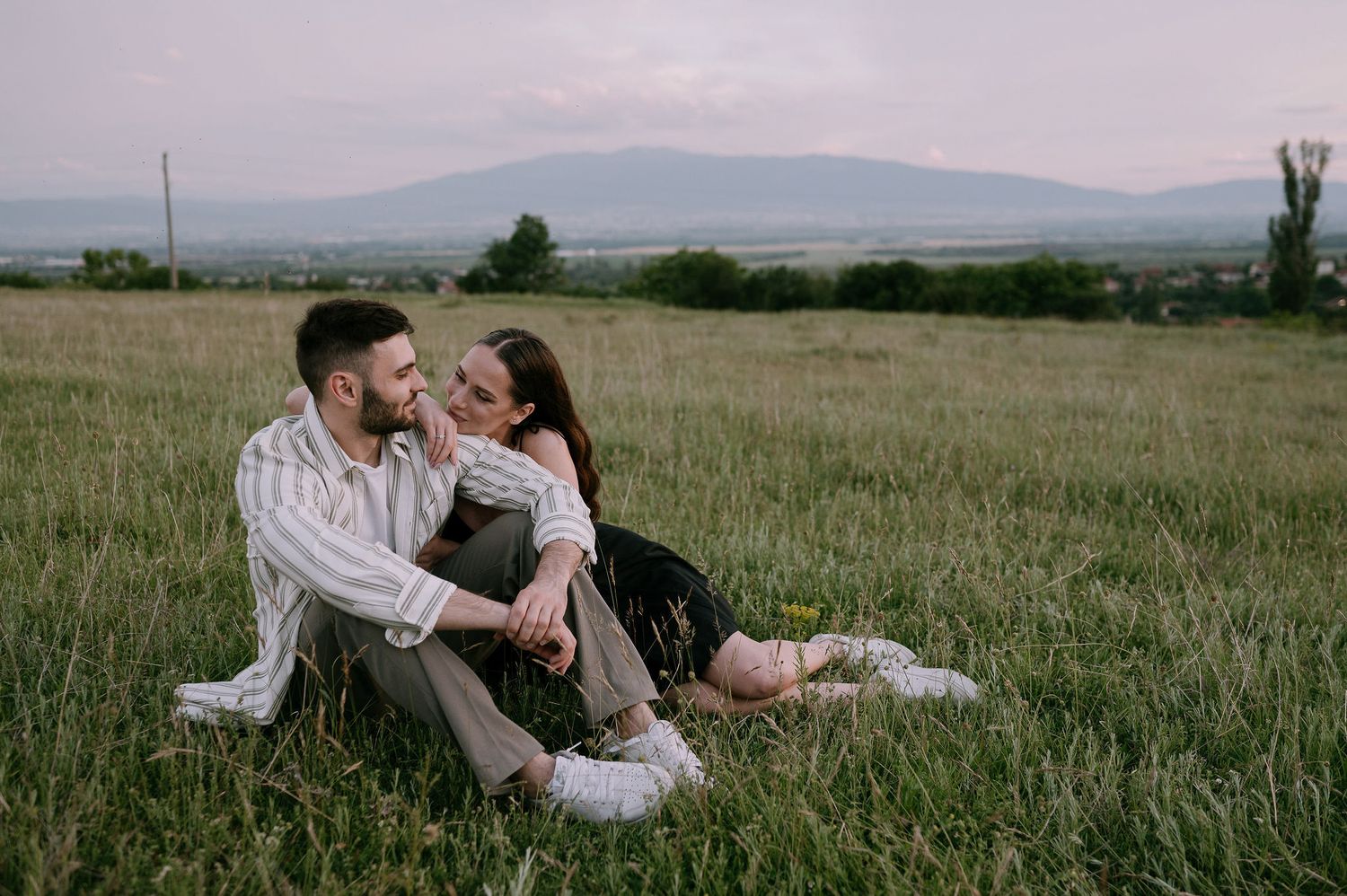 Couple sharing tender moments together while lying in a grassy meadow with mountains in the background.