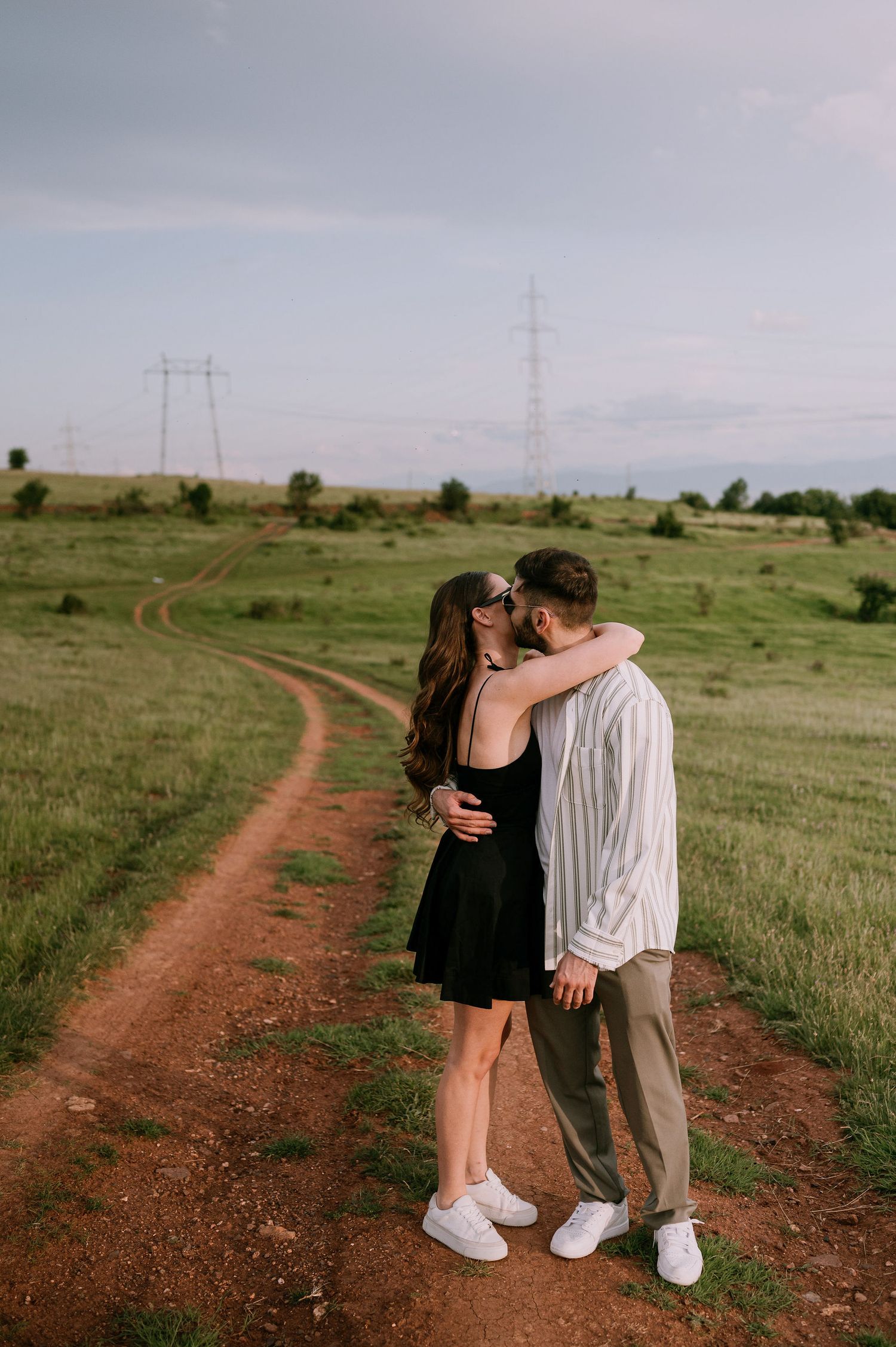 A couple shares romantic moments along a winding dirt path through green fields with power lines in the distance.