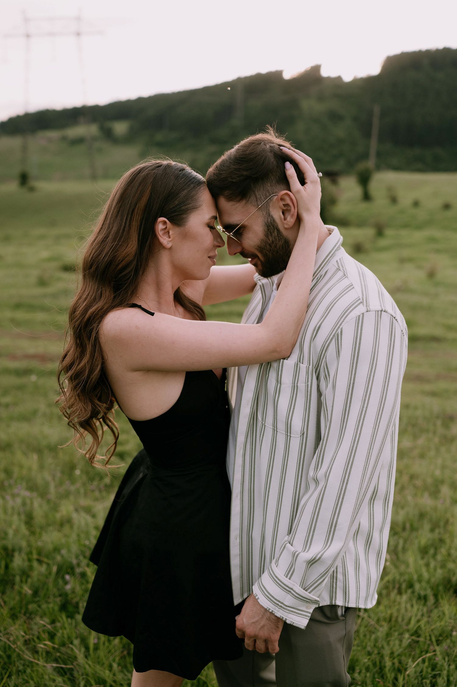 A romantic couple embraces in a green meadow at sunset, with her in a black dress and him in a striped shirt.