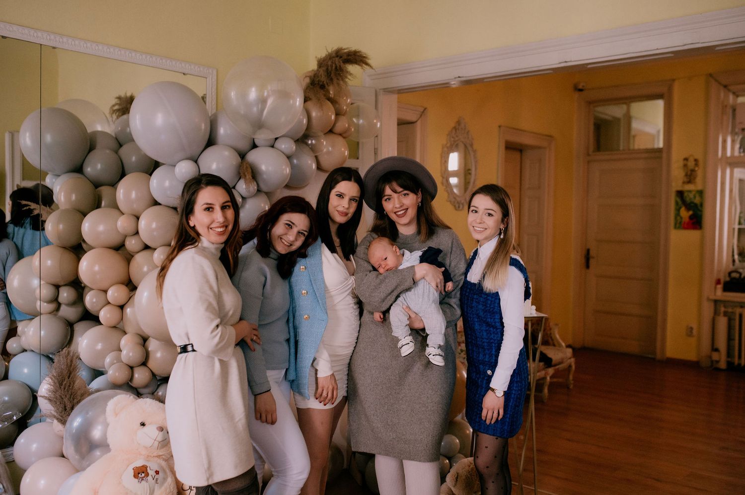 A group of people gather in a hallway decorated with gray and white balloons for a celebration.