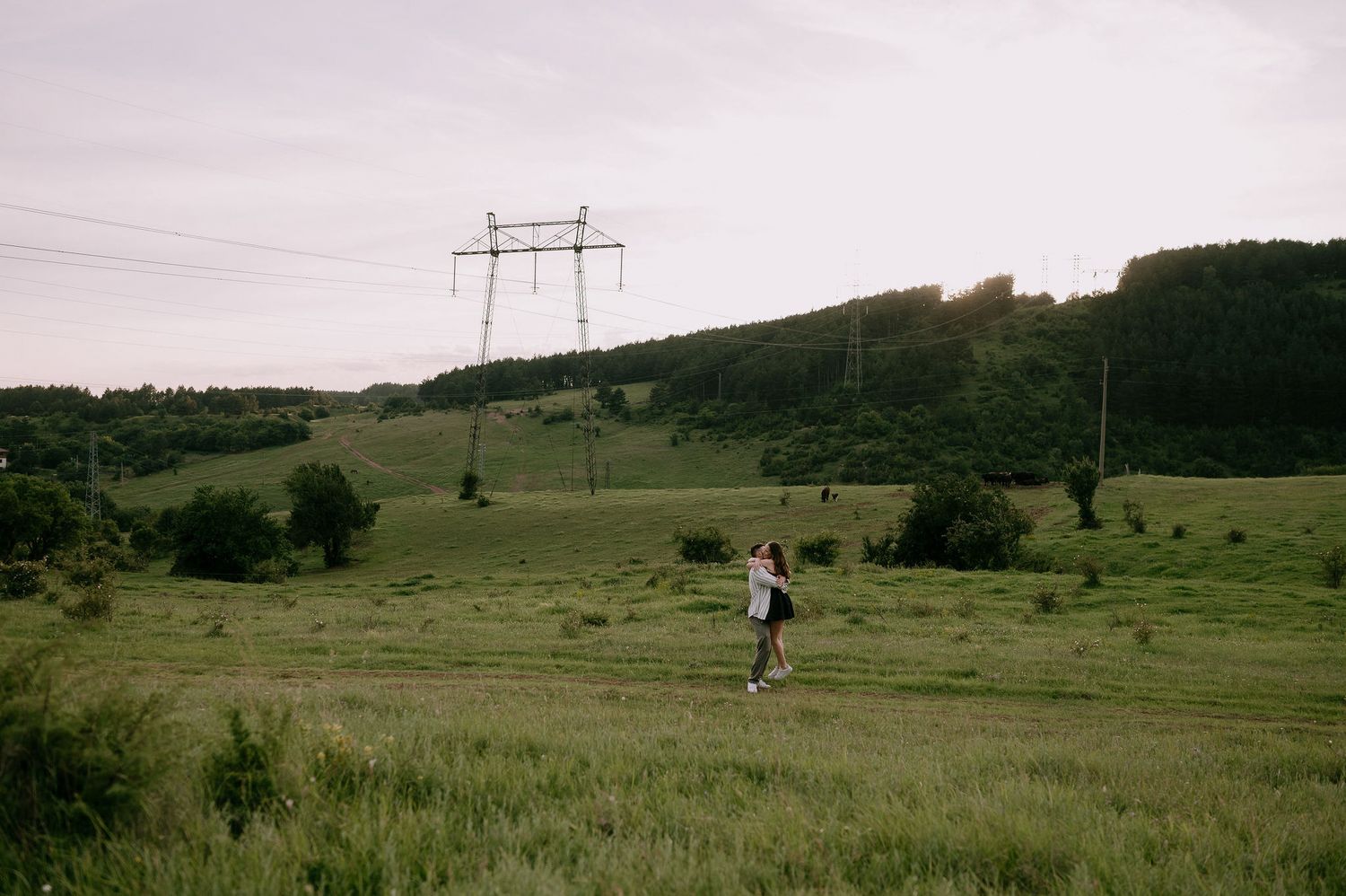 People walk through a scenic green meadow with power lines and rolling hills in the distance on a cloudy day.