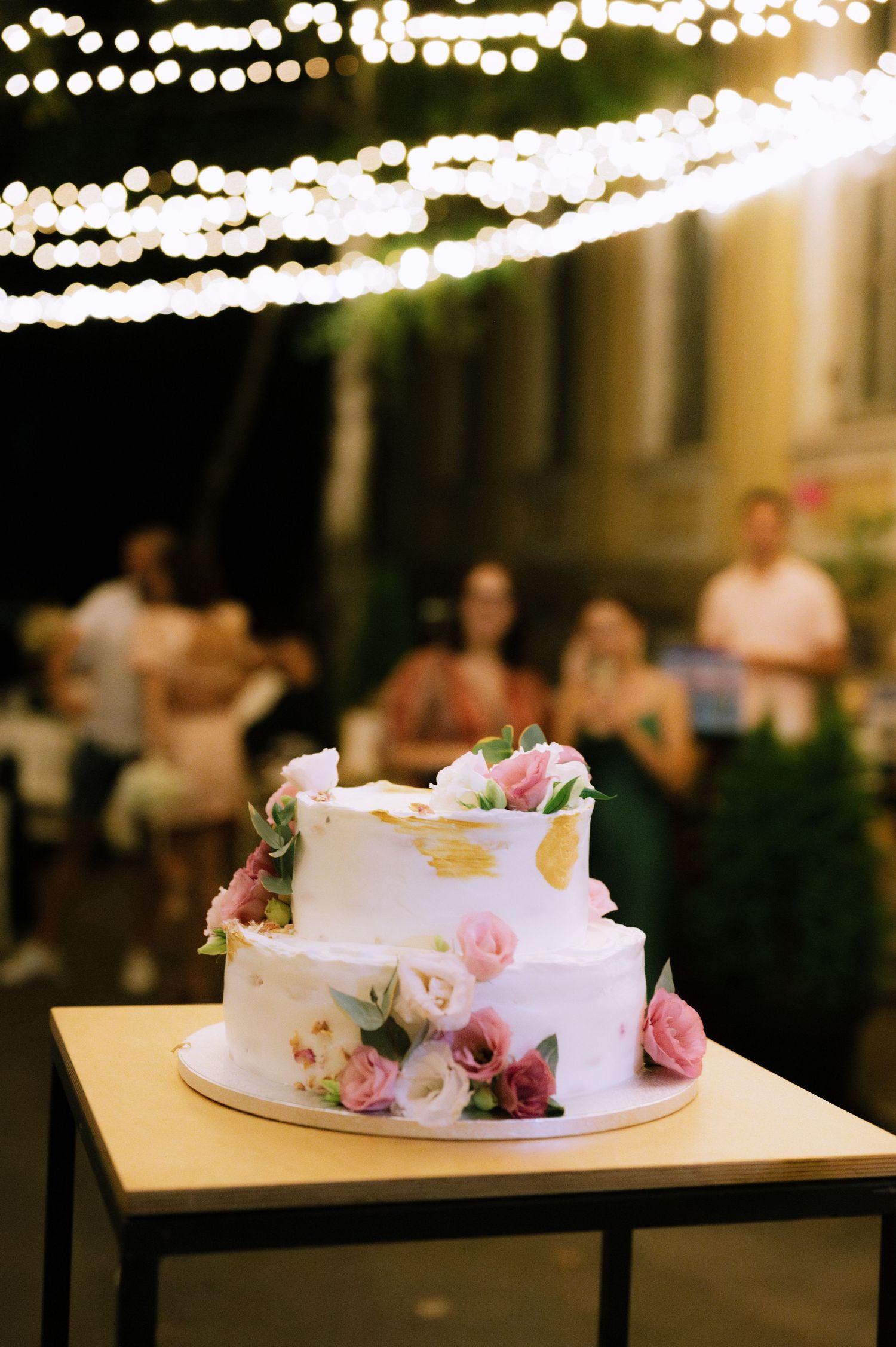 Two-tiered white wedding cake decorated with pink and burgundy flowers sits on table with string lights in background.