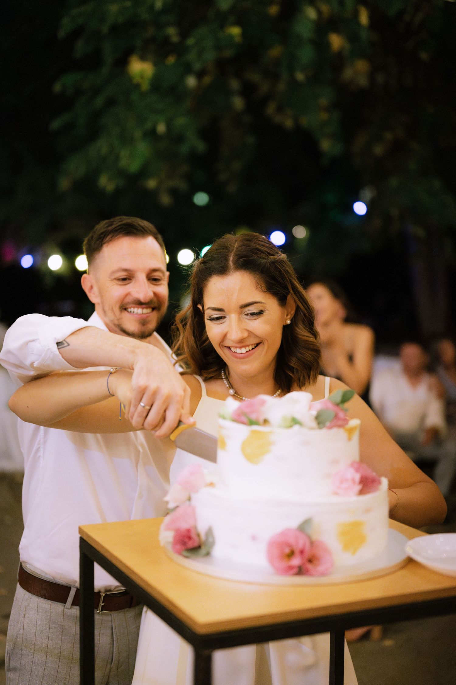 A couple shares a sweet moment cutting their wedding cake decorated with pink flowers at an outdoor evening reception.