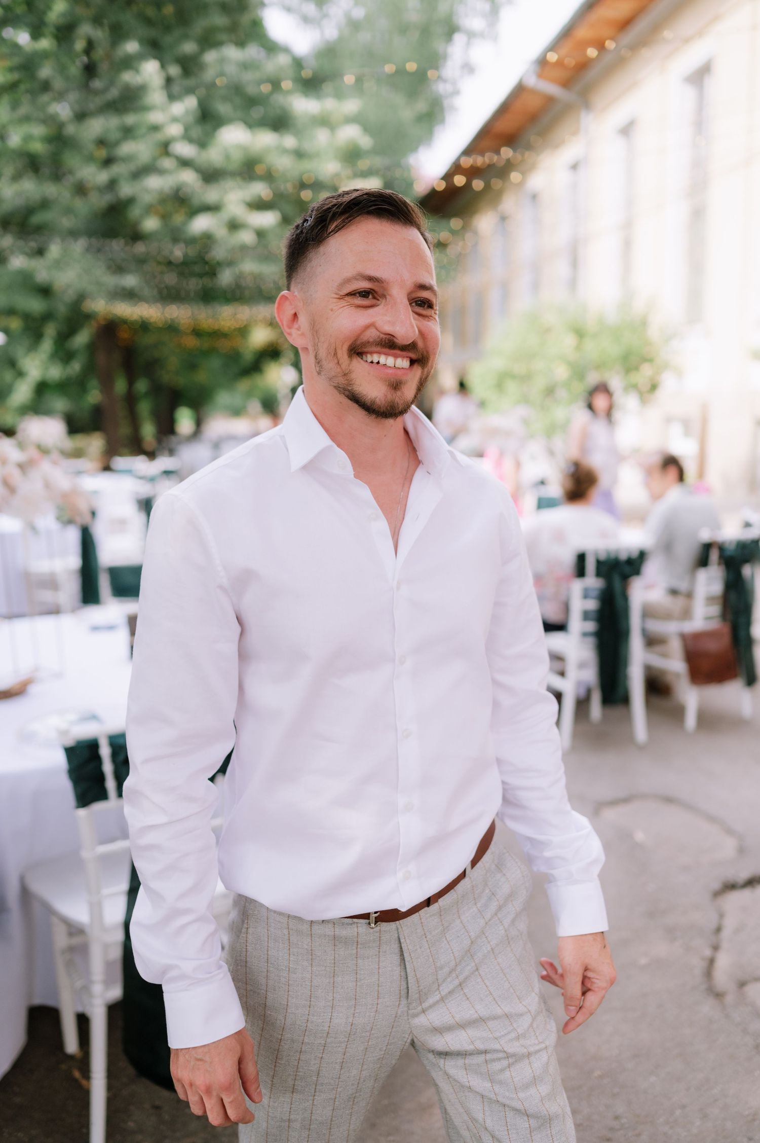 A person in a crisp white dress shirt and light gray pants smiles at an outdoor event venue.