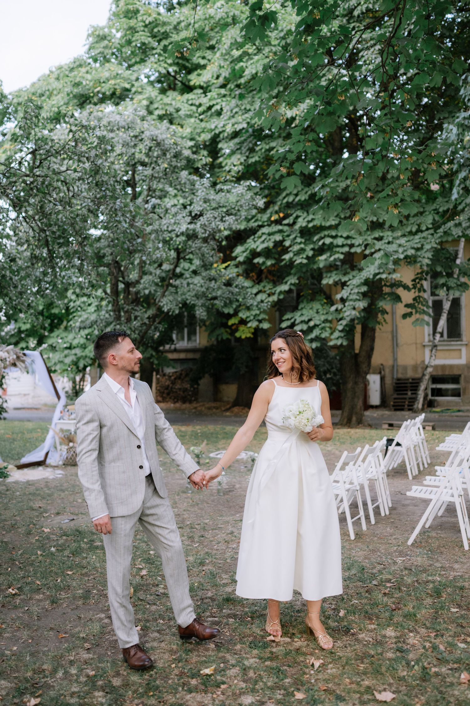 A couple in wedding attire dance and twirl playfully on a lawn surrounded by white chairs and green trees.