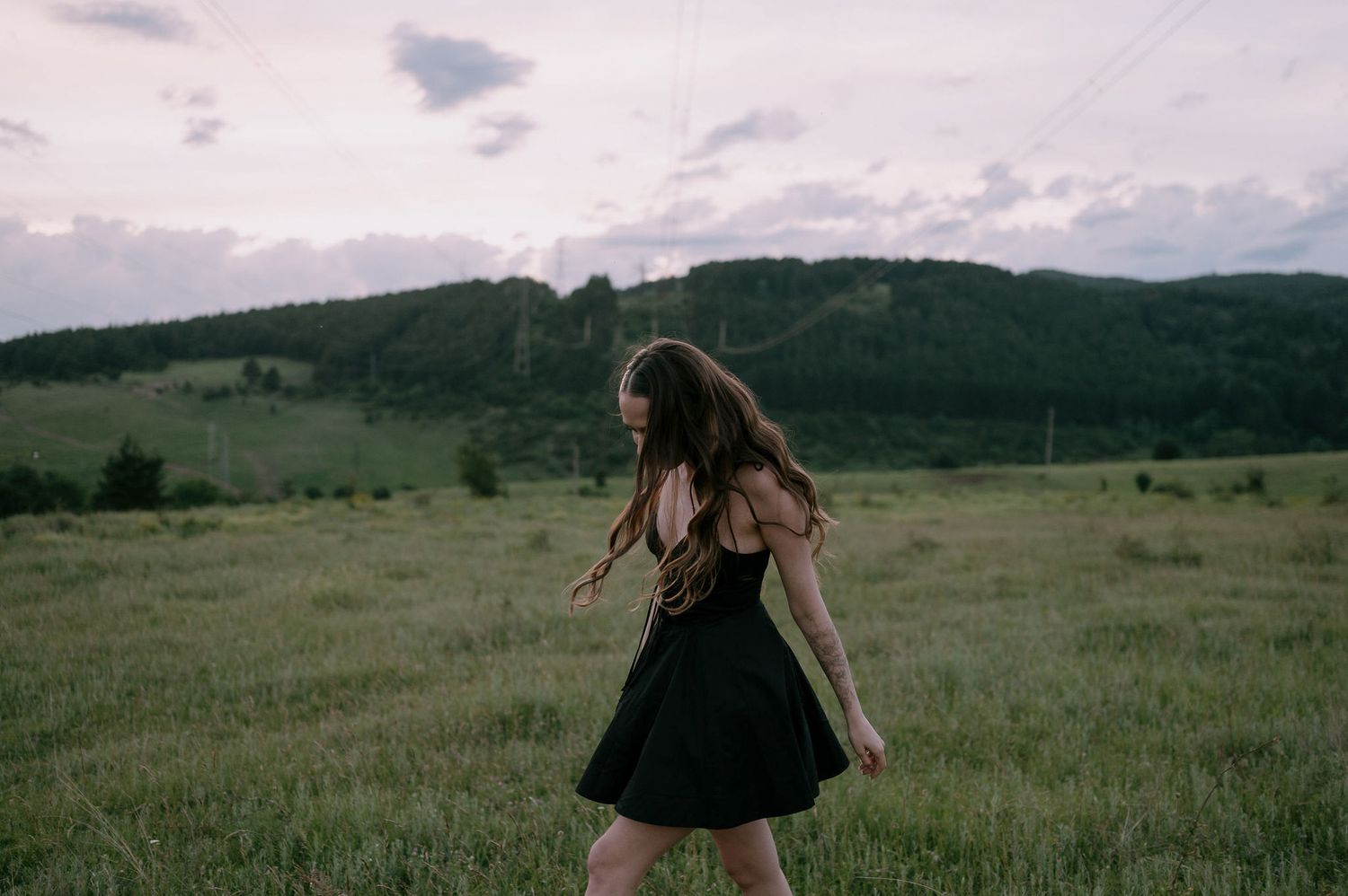 A person in a black dress twirls gracefully in a scenic meadow with rolling hills and cloudy skies in the background.