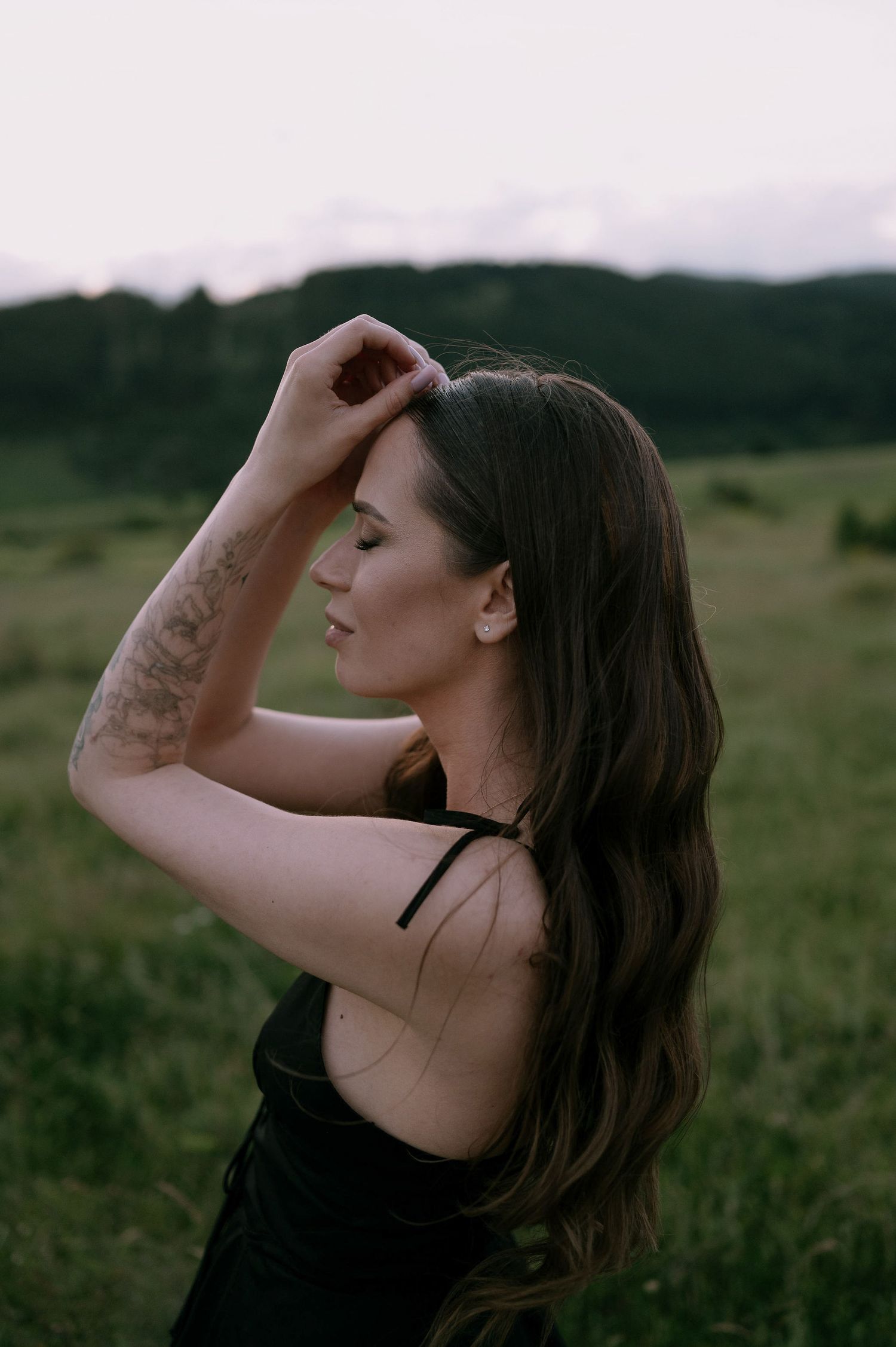 A person stands in profile against a moody natural landscape with their hand raised to their forehead at sunset.