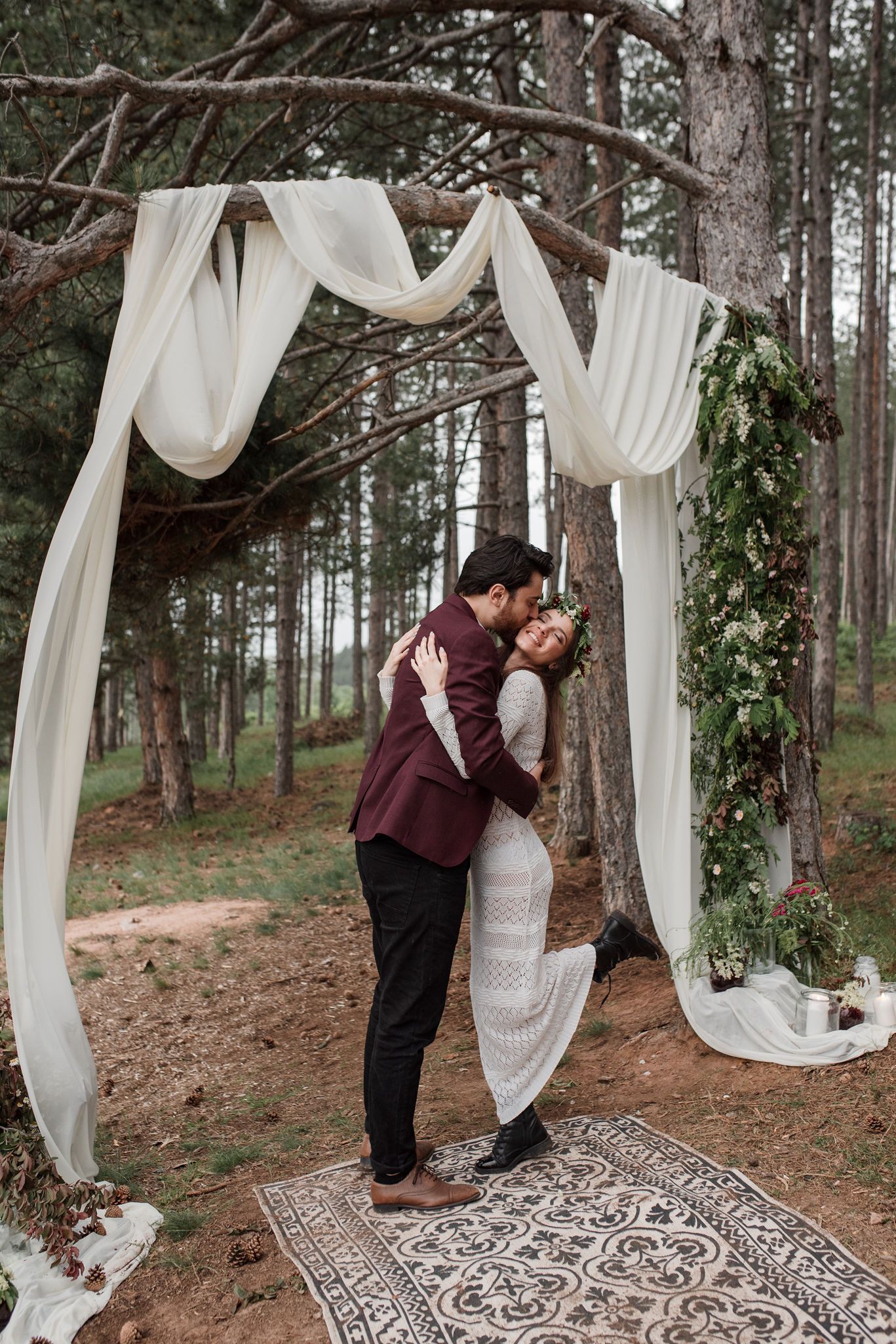 A romantic outdoor wedding ceremony setup in a forest with white draping fabric and a decorative rug on the ground.