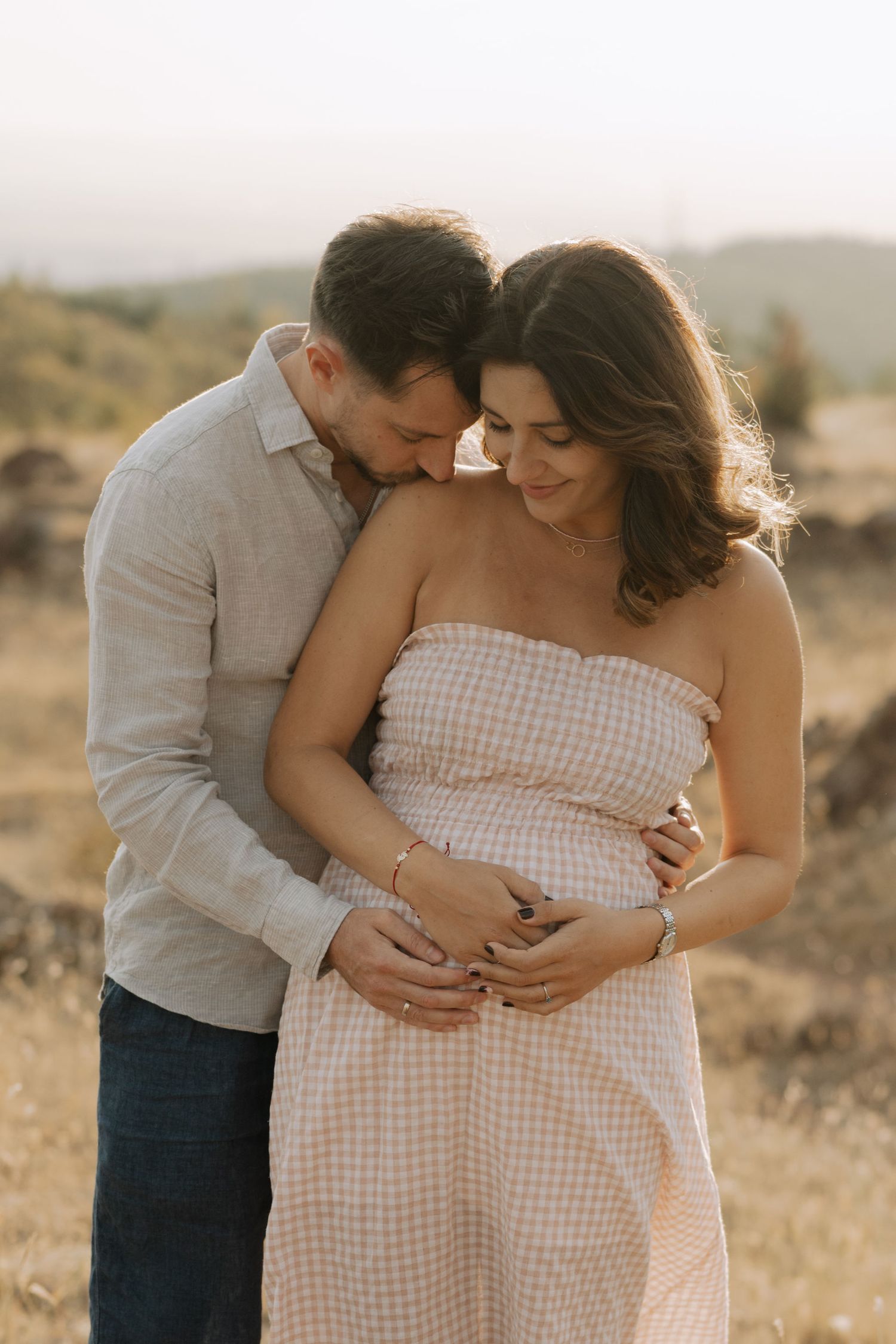 Expectant couple embraces tenderly in a sun-drenched desert landscape wearing white and grey casual attire.