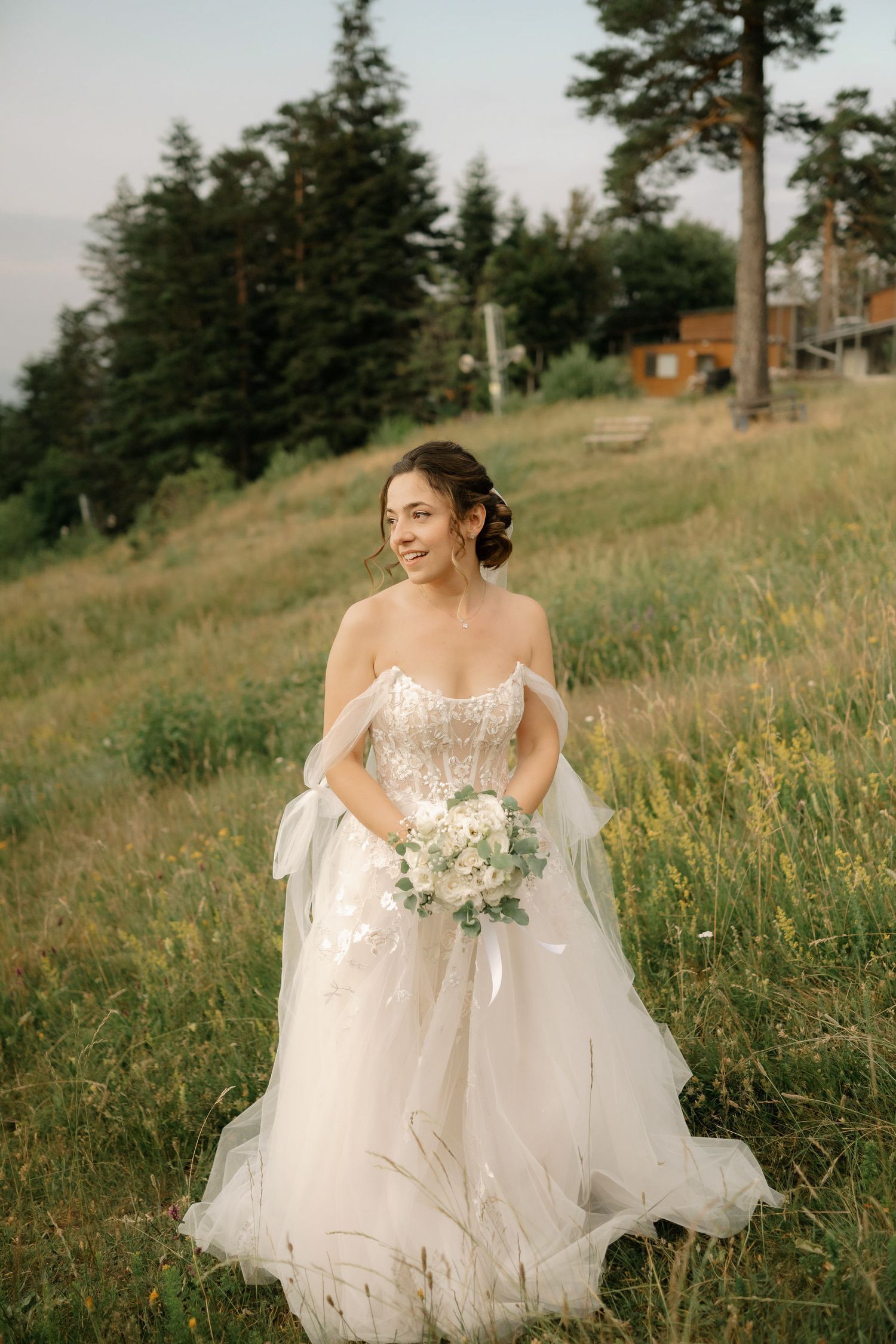 A bride in a flowing white strapless gown holds a bouquet while standing on a grassy hillside with evergreen trees.
