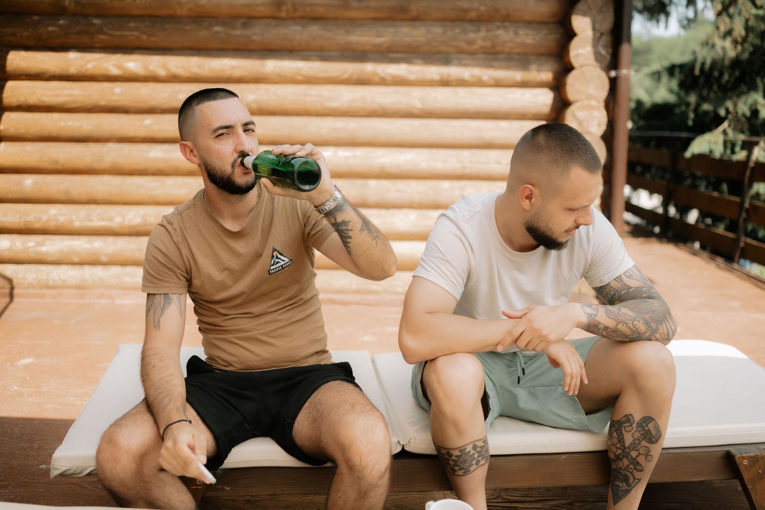 Group of friends sitting on wooden bench outdoors drinking beer and socializing together.